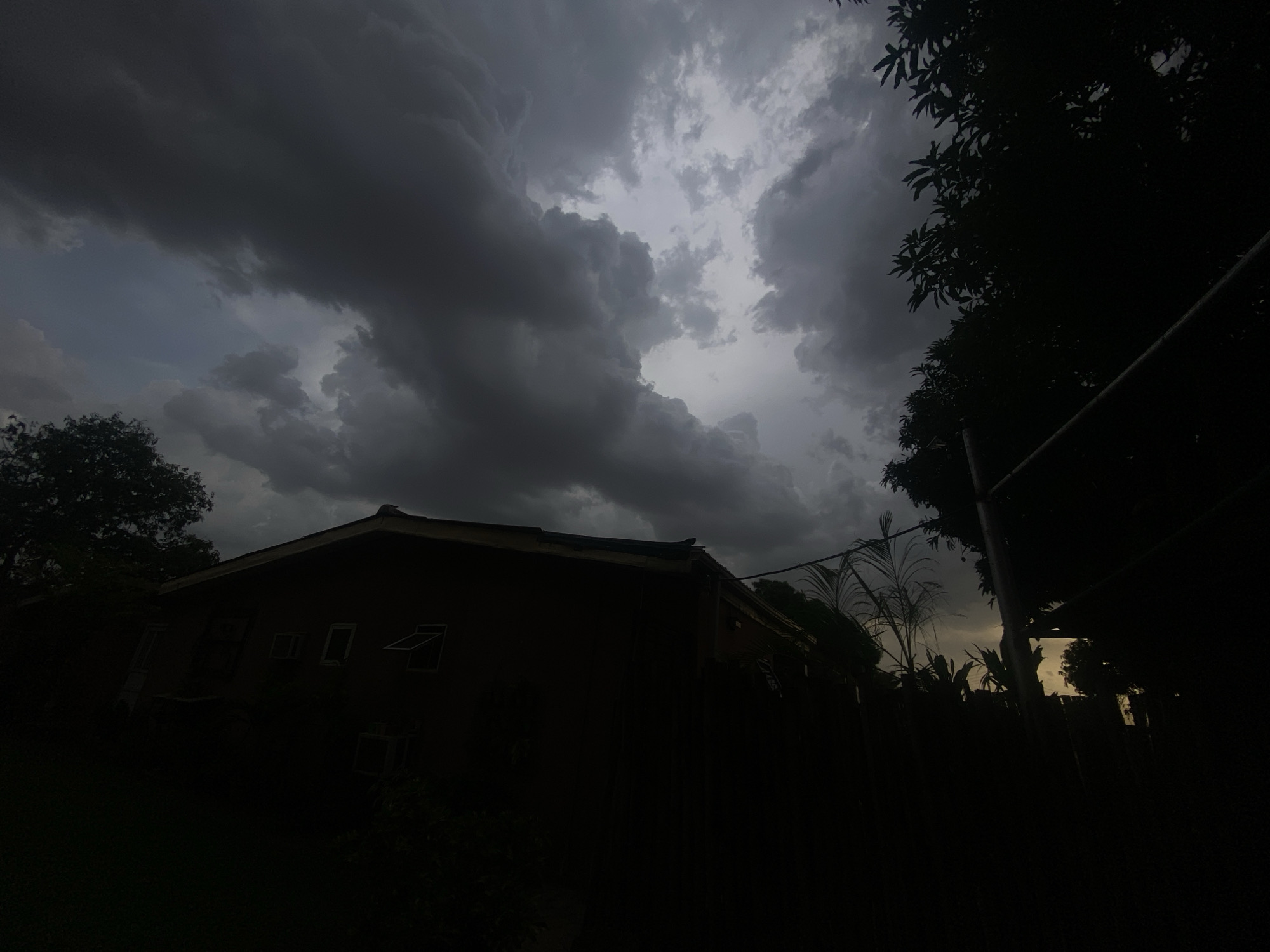 The image depicts a dimly lit scene in Barden Yana, Nigeria, under a heavy, overcast sky filled with dark, ominous clouds. A single-story building is silhouetted against the sky, appearing dark with only the outlines of windows visible. Trees and foliage surround the building, adding to the darkness of the foreground. A pole and some indistinct structures are visible on the right side of the frame, further obscuring the details of the surroundings. The light is faint, casting long shadows and making it difficult to discern specific details.