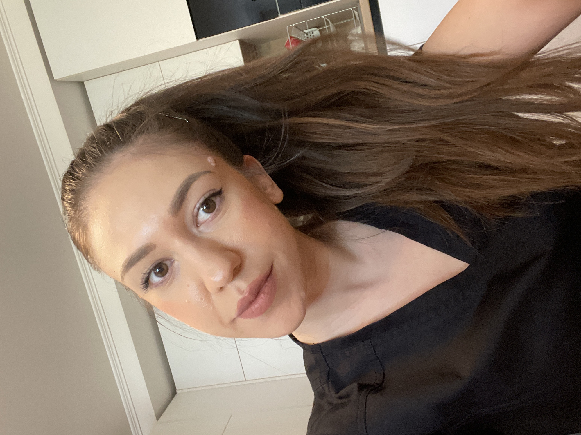 A woman with long, brown hair, light brown eyes, and a fair complexion is facing the camera. She is wearing a black garment. The background includes what appears to be part of a room with light-colored cabinets and shelves. There is no clear indication of Sofia, Bulgaria in this image. The image appears to be a selfie.