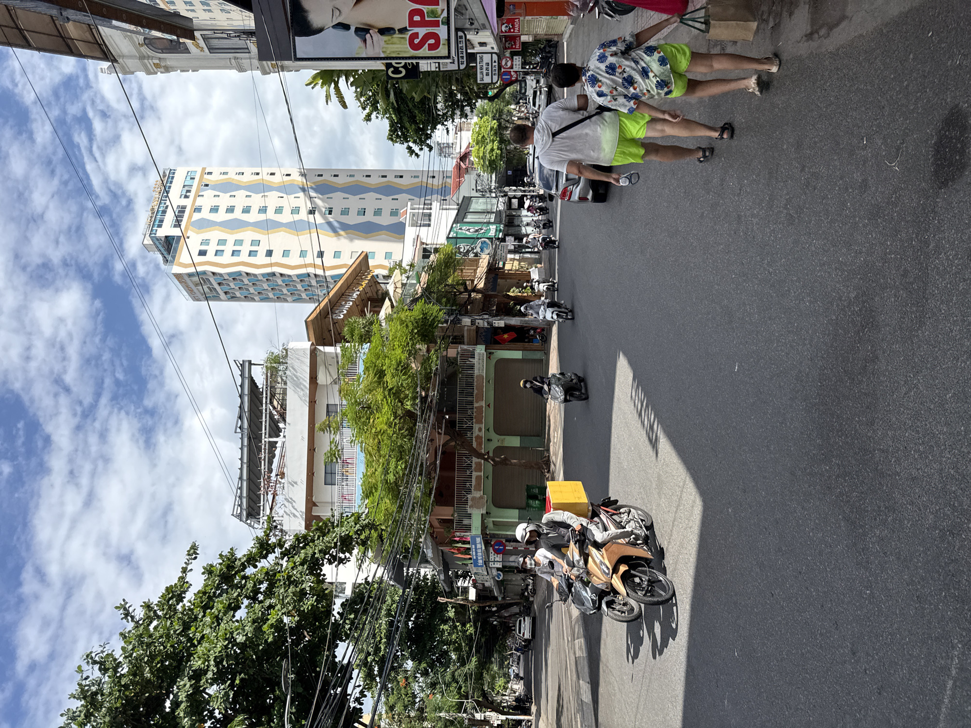 In Nha Trang, Vietnam, a street scene unfolds under a partly cloudy sky. The road is paved, with a distinct shadow cast from the right side, indicating a bright sun. On the left side of the street, lush green trees line the sidewalks. Buildings of varying heights and architectural styles dominate the background, including a modern, tall building with a distinctive blue and gold zig-zag pattern and a smaller, white building with balconies. Several streetlights and power lines crisscross the sky above.
The street is active with traffic, primarily motorbikes. Two motorbikes are in the foreground, one gold and one black, with riders wearing helmets and casual clothing. The gold motorbike has a large yellow box attached to its rear. Further down the street, more motorbikes are visible, some with passengers. Two people, a man and a child, walk away from the camera on the right sidewalk, both wearing bright green shorts. The man wears a white t-shirt and sandals, while the child wears a floral-print shirt. A shopping bag is visible in the child's hand. The sidewalks are lined with shops and businesses, indicated by signage and storefronts. The overall atmosphere suggests a typical day in a bustling Vietnamese city.