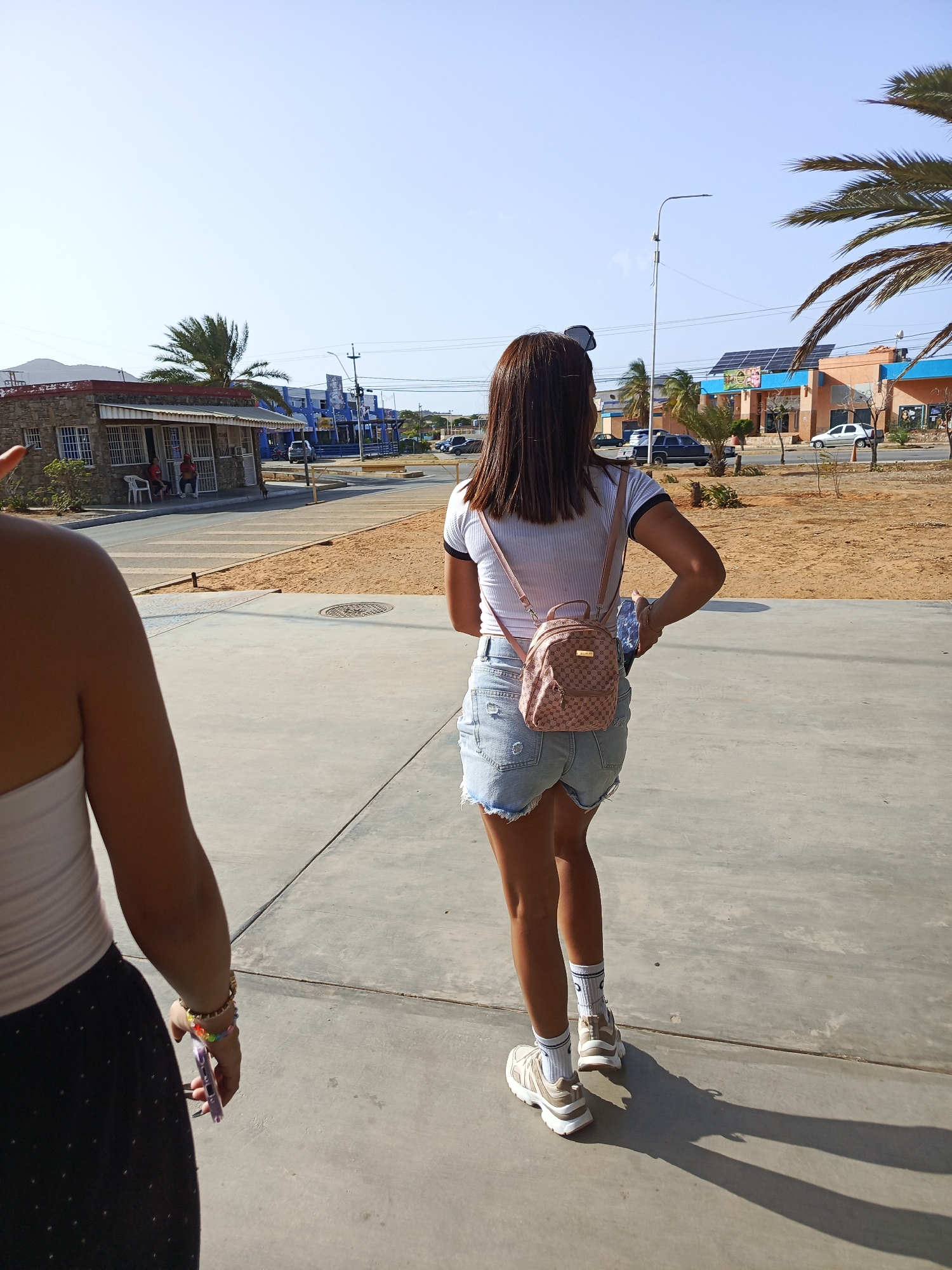 This outdoor image captures two women walking along a concrete sidewalk on a bright, sunny day in Juan Griego, Venezuela. The time of day is indicated by the harsh shadows cast by the subjects and objects, suggesting midday or early afternoon with a clear sky.
In the foreground on the left, a woman is partially visible from the chest down, wearing a white sleeveless top and a dark skirt or trousers with a subtle pattern. She is holding a light purple smartphone in her right hand and wears multiple bracelets, including colorful beaded ones and a gold-toned one, on her wrist.
The central subject is a woman walking away from the camera. She wears a white t-shirt with black trim on the sleeves, light blue distressed denim shorts, white athletic socks with blue patterns, and light-colored athletic shoes. A small pink backpack with a repeating pattern, possibly a logo, is worn on her back. She also has sunglasses perched on her head.
The background reveals a typical street scene. To the left, there's a low-rise building with a stone facade and a red roof, featuring barred windows and an awning. People are visible sitting outside this building. Further down the street, various other buildings are present, including one painted blue and another with an orange and blue facade, which has solar panels installed on its roof. Palm trees are scattered throughout the scene, along with utility poles, power lines, and a street lamp. Several cars are parked or driving on the paved road. The ground bordering the sidewalk and road is sandy and sparse, typical of an arid or coastal environment. The overall atmosphere is casual and relaxed. No clearly legible text or brands are visible, apart from the repetitive pattern on the backpack which resembles a logo.