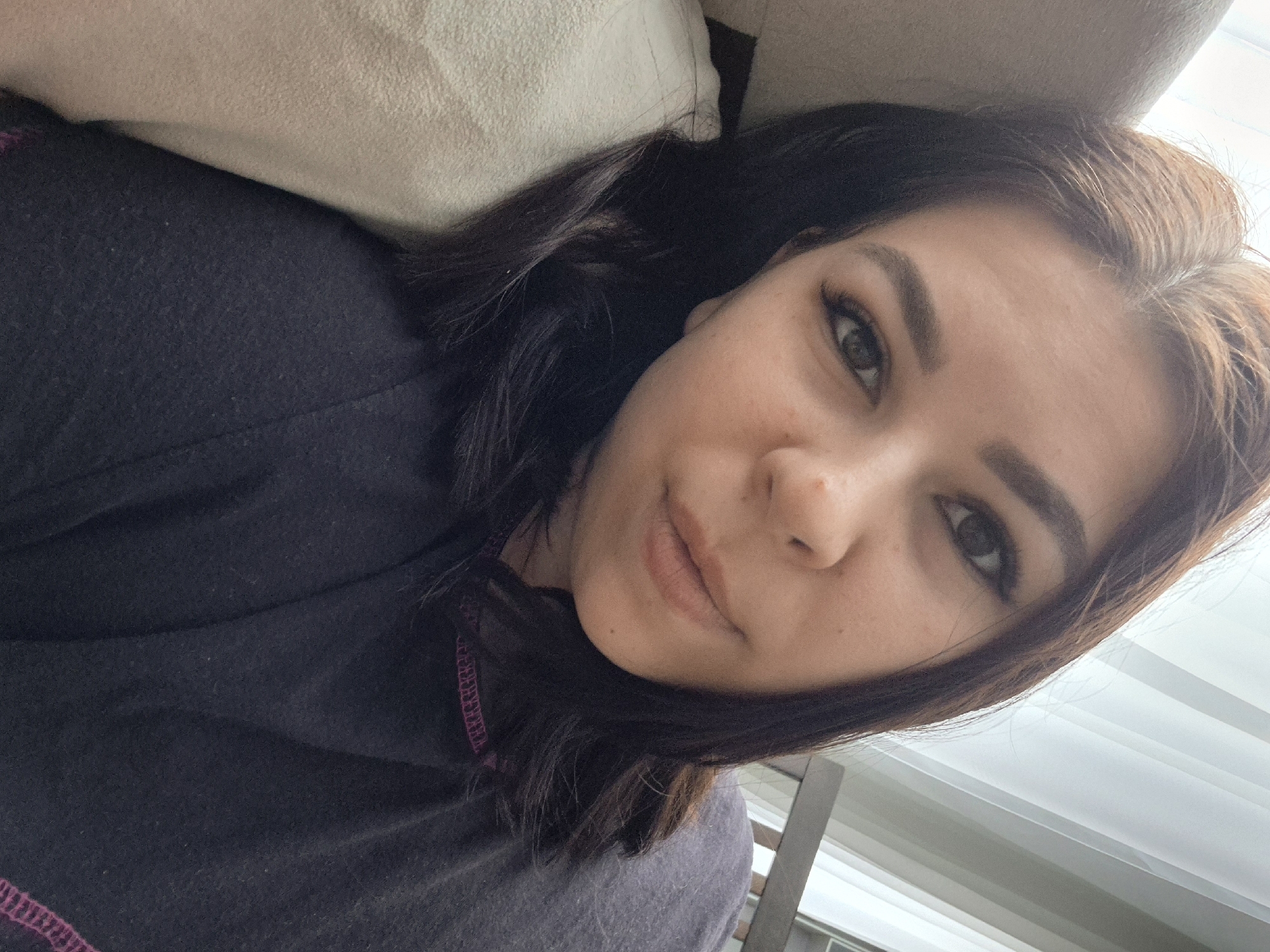 A woman lies on a light-colored cushion, wearing a dark gray top with purple stitching around the neckline. Her dark brown hair is parted and falls around her face. She looks directly at the camera with a neutral expression. The background includes a partially visible window with white, vertical blinds or panels. The location cannot be determined based on the image.