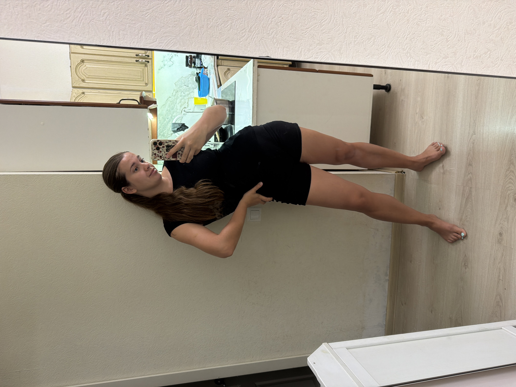 The image shows a woman with long brown hair pulled back in a ponytail, wearing a black t-shirt and black shorts. She is standing in what appears to be a kitchen and is taking a mirror selfie with her phone. The kitchen has white cabinets and a white countertop. There is a sink and a stovetop visible. The floor is light wood. The woman's left hand is on her hip, and her right arm is extended, holding the phone. Her feet are bare, and her toenails are painted a light blue. The overall lighting is neutral. The image does not provide any specific visual cues to determine the exact location within Sochi, Russia.