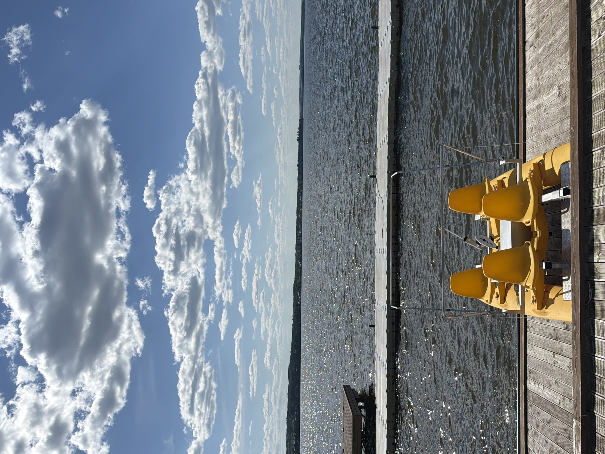 This image captures a serene waterside scene in Votkinsk, Russia, under a bright, sunny sky. The upper portion of the frame is dominated by a clear blue sky, interspersed with numerous white cumulus clouds of varying sizes and densities; some are large and shadowy, while others are smaller and more defined.
The midground features a wide expanse of water, likely a lake, which intensely reflects sunlight, creating countless sparkling glints across its rippled surface. In the foreground, a weathered wooden dock extends horizontally across the bottom of the image, where a bright yellow four-seater pedal boat is moored. The boat is seen from the rear, prominently displaying its four individual yellow bucket seats, arranged in two rows, along with a metal frame and what appear to be steering and pedaling mechanisms.
Immediately beyond the pedal boat, a grey modular floating dock stretches horizontally across the water. To the left, a smaller, darker wooden platform is also visible on the water. In the distant background, a low, tree-lined shoreline, dotted with faint structural outlines, defines the horizon under the bright conditions. There are no individuals present in the scene, and no explicit actions or interactions are depicted. The overall environment suggests a recreational area on a clear, warm day.