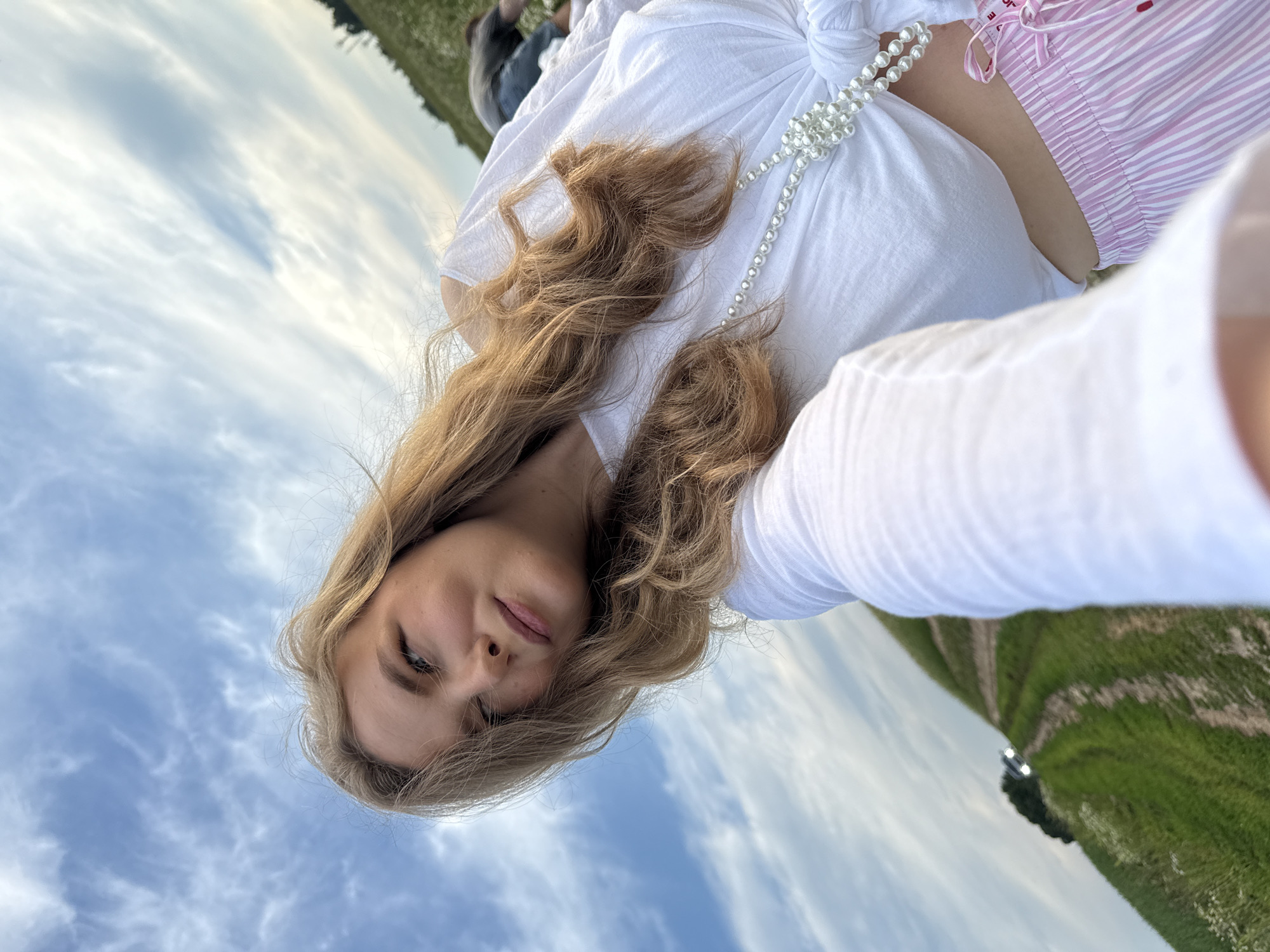 A young woman with long blonde hair is taking a selfie outdoors. She is wearing a white knotted top, pink striped pants, and a pearl necklace. The background shows a field with a path leading up a hill, a car in the distance, and a cloudy blue sky. Another person is visible in the background lying in the field. The location appears to be a rural area.