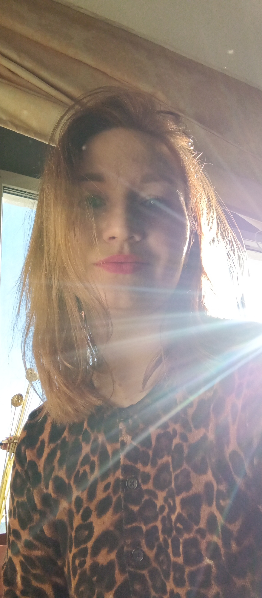 A photograph shows a woman from a low angle. She has auburn hair, pink lipstick, and wears a leopard print cardigan. Sunlight streams in from the right, partially obscuring her face. A window is visible to the left, showing a blue sky and an object resembling a Ferris wheel. The background includes what appears to be beige drapery. Location: Unknown, Unknown.