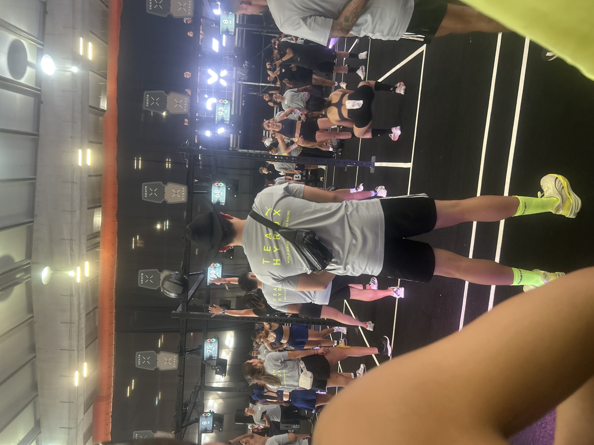 This image shows an indoor event with many people in an exercise studio. There are exercise stations set up throughout the room. The event is likely related to the "HYROX" brand, as their logo is displayed on multiple screens and on the participants' clothing. The people appear to be engaged in some kind of fitness activity, possibly a group workout or competition. The scene is busy with people in workout attire and athletic shoes. There are no specific landmarks indicating the location in the city of Unknown, Unknown.