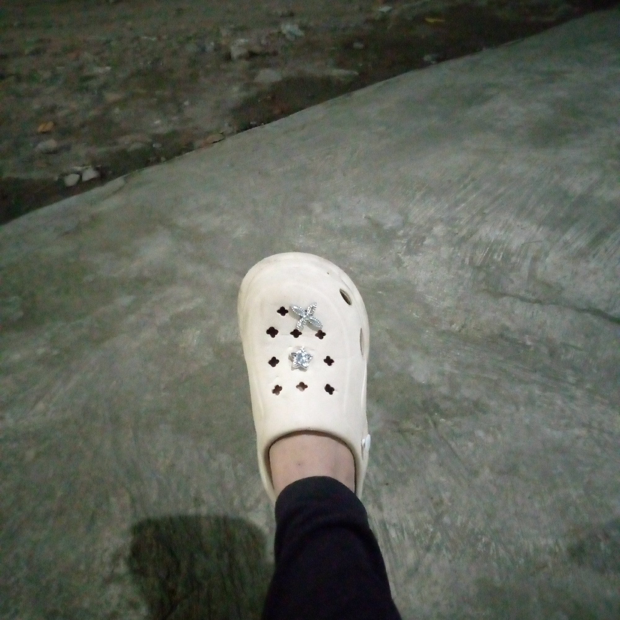 A low-angle, medium shot shows a cream-colored Croc shoe with two charms on it, a silver butterfly and a silver star with a clear gem, on a person's foot. The foot is clad in black pants. The shoe rests on a mottled gray concrete surface, which appears to be outdoors. Behind the concrete area, a rough, darker surface with some debris is visible, possibly indicating a roadside or an unpaved area. The overall lighting suggests it is nighttime or dusk. The scene is located in Sumberwono, Indonesia.