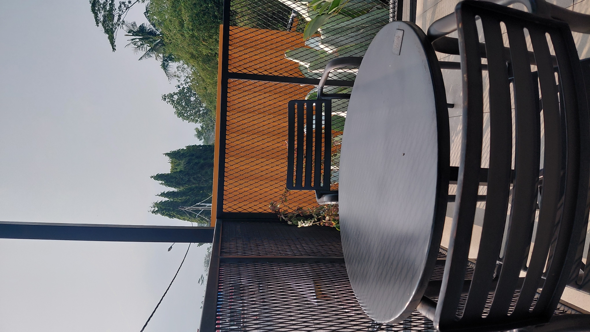An outdoor seating area with a round black table and two black chairs is shown in this image. The table is in the foreground, and a chair is visible behind it, facing away from the camera. Another chair is partially visible on the right side of the image, also facing away. The seating area is enclosed by a black metal mesh fence. Behind the fence, there is an orange wall with a diamond pattern. In the background, several tall, green trees are visible against a pale sky. The overall scene suggests a calm, outdoor dining or relaxation space. The location is identified as Karanglewas, Indonesia.