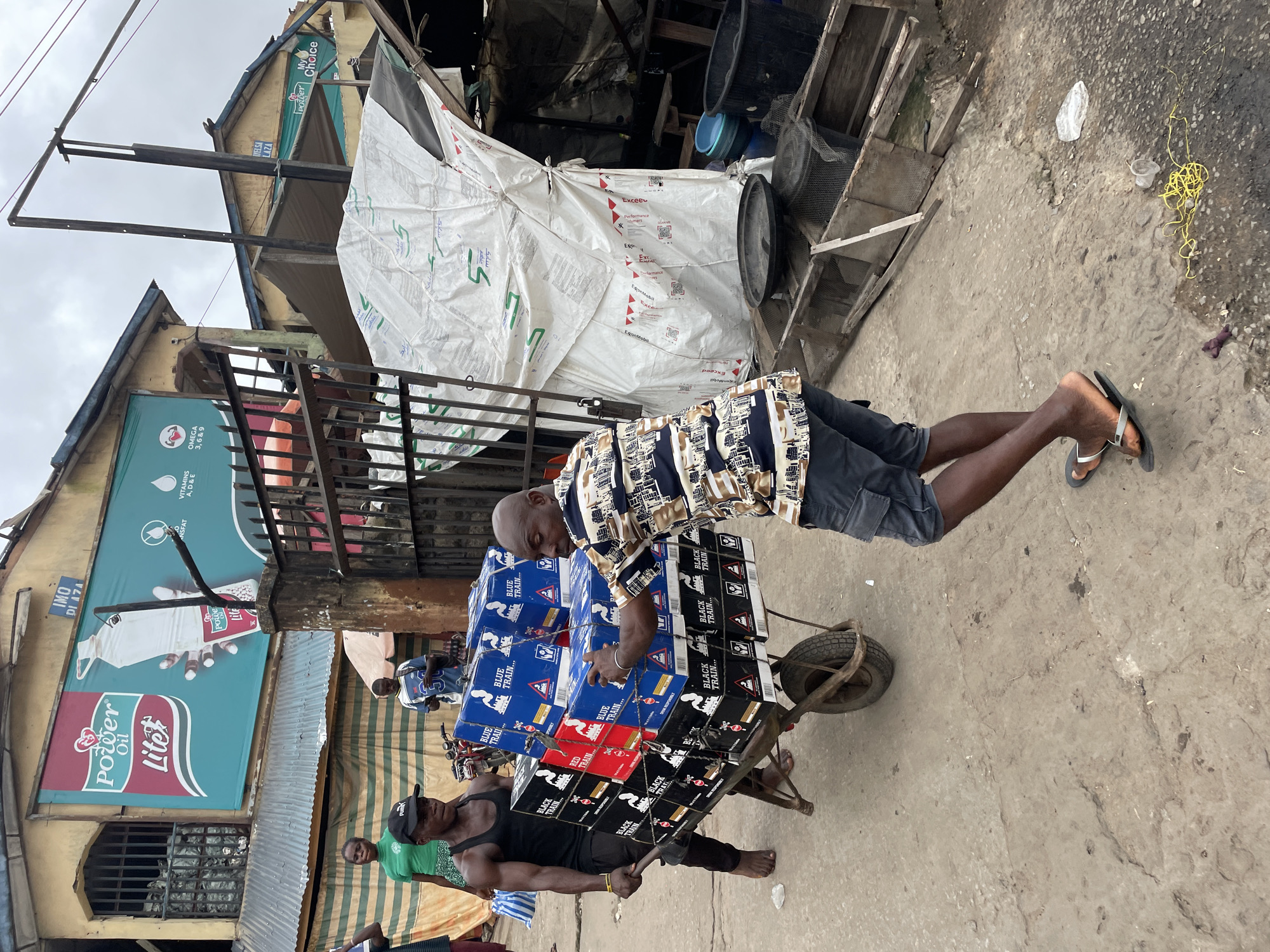 In Unknown, Unknown, two men are actively engaged in transporting a heavily loaded wheelbarrow filled with boxes. The man on the left, wearing a black tank top and black pants, is pushing the wheelbarrow. He is barefoot and has a muscular build. The man on the right, dressed in a patterned short-sleeved shirt and grey shorts, is assisting by steadying the load. He is wearing sandals. The wheelbarrow contains numerous stacked boxes, some labeled "BLUE TRAIN" and others "BLACK TRAIN." The scene appears to be outdoors, on a paved surface, with buildings and market stalls visible in the background. The sky is overcast.