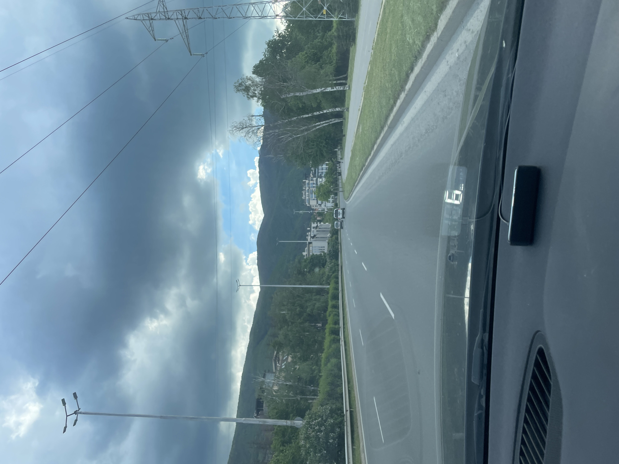A road stretches forward, viewed from inside a vehicle. The road, marked with white lines, leads towards a distant mountain range. Trees line the roadside. Overhead power lines are visible against a cloudy sky. In the vehicle, a digital display shows the number "69". A black object is affixed to the dashboard. The location is Unknown, Unknown.
