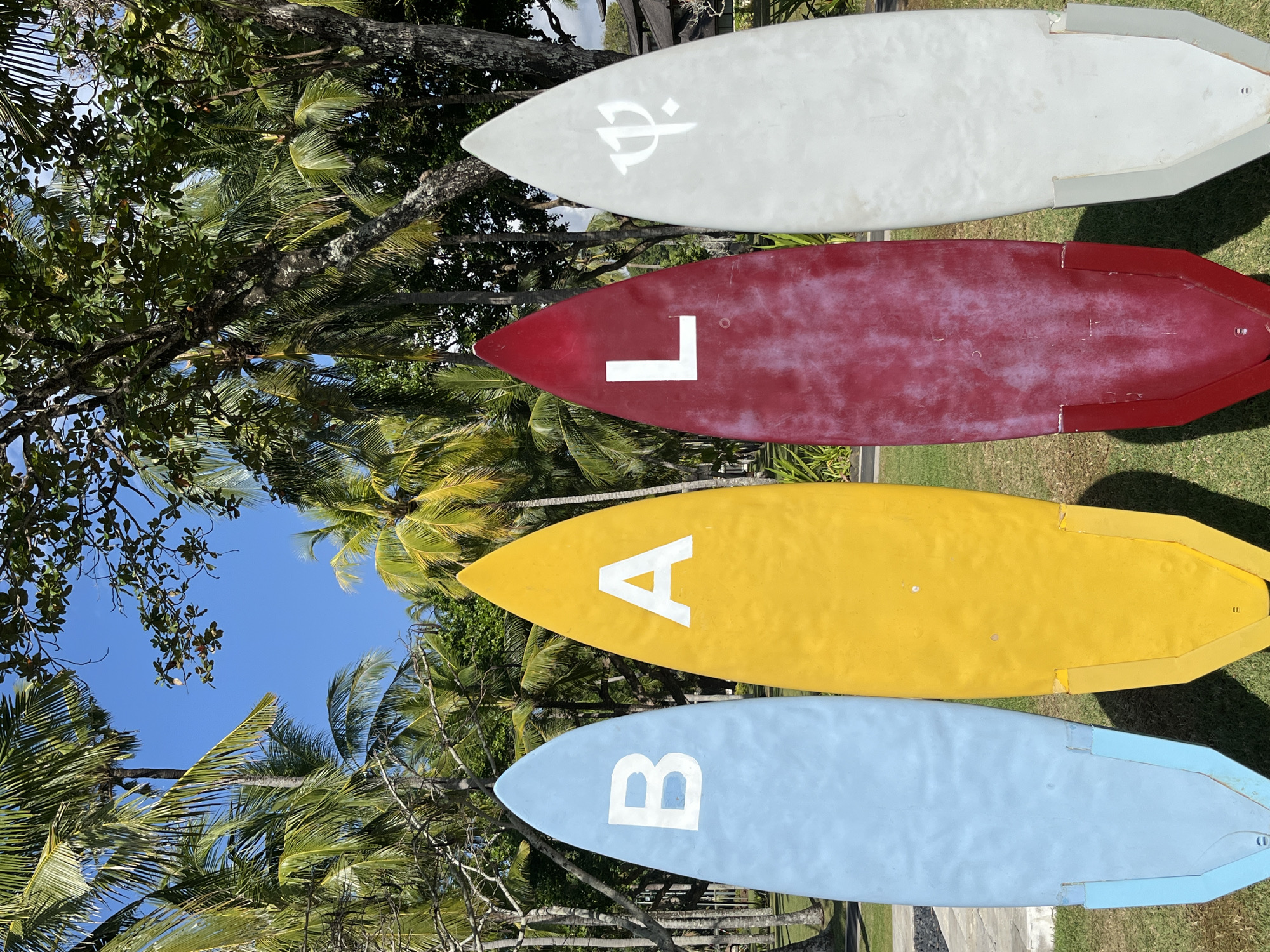 In Kuta Selatan, Indonesia, four surfboards are arranged side-by-side on a grassy area, facing the viewer. The surfboards are in distinct colors: light blue, yellow, red, and white. Each of the first three surfboards displays a large, white uppercase letter: "B" on the blue board, "A" on the yellow board, and "L" on the red board. The white surfboard features a white symbol resembling a trident with a circle at the base. The surfboards are positioned upright, seemingly as a decorative display, with their bottoms resting on the grass. Behind the surfboards, a backdrop of lush green palm trees and foliage under a clear blue sky suggests a tropical outdoor environment. Some trees have visible branches and leaves, and the sunlight indicates it is daytime. The overall scene is bright and sunny, with a hint of wear and tear visible on the surfaces of the red and white surfboards.