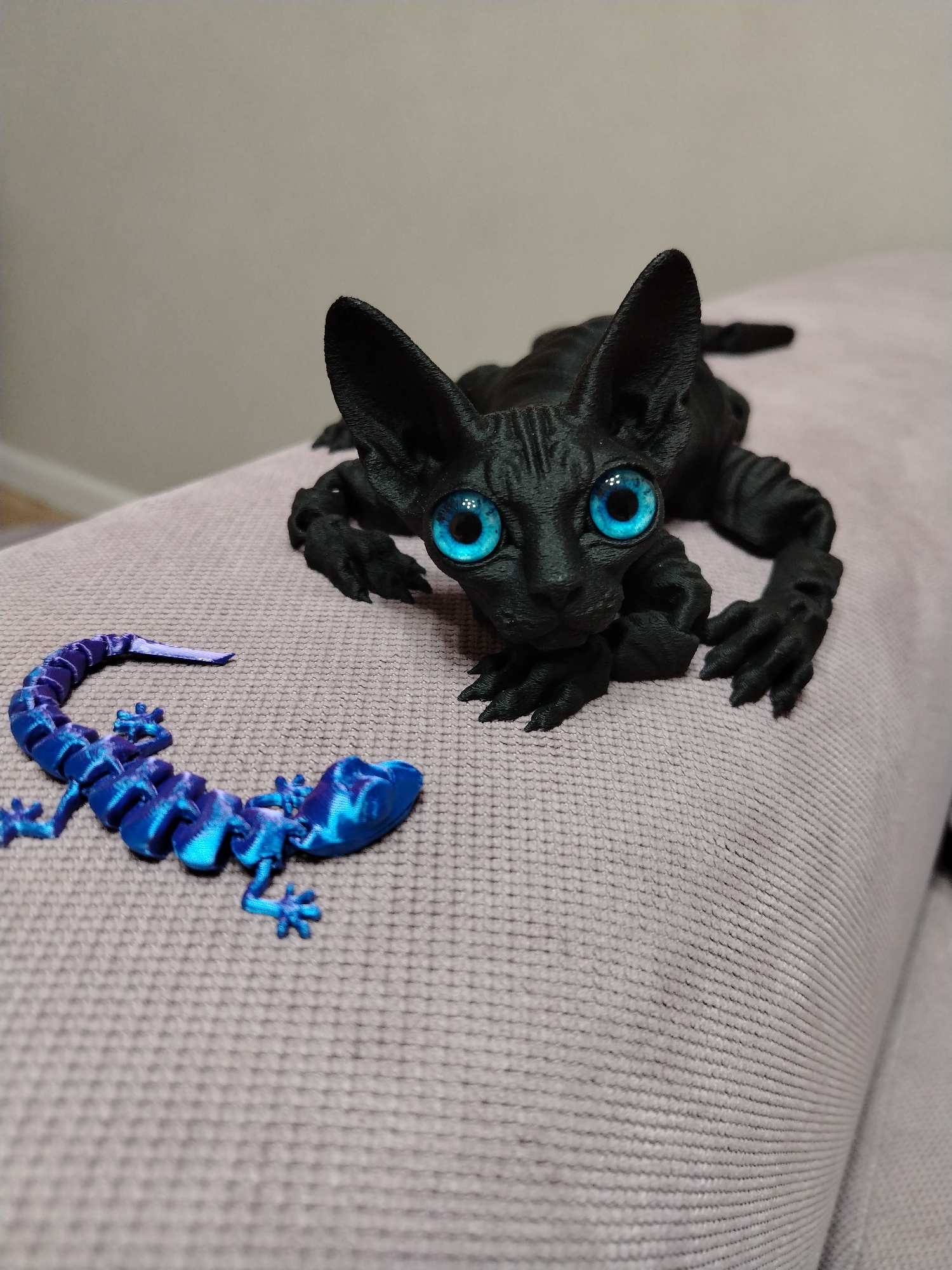 The image shows two 3D-printed figures. The first figure is a black cat with large, bright blue eyes. It is posed on a light-purple upholstered surface, likely a sofa or chair. The second figure, located to the left of the cat, is a blue and purple articulated lizard. The background is a blurry, light-colored wall. There is no indication of the location being in Izhevsk, Russia.