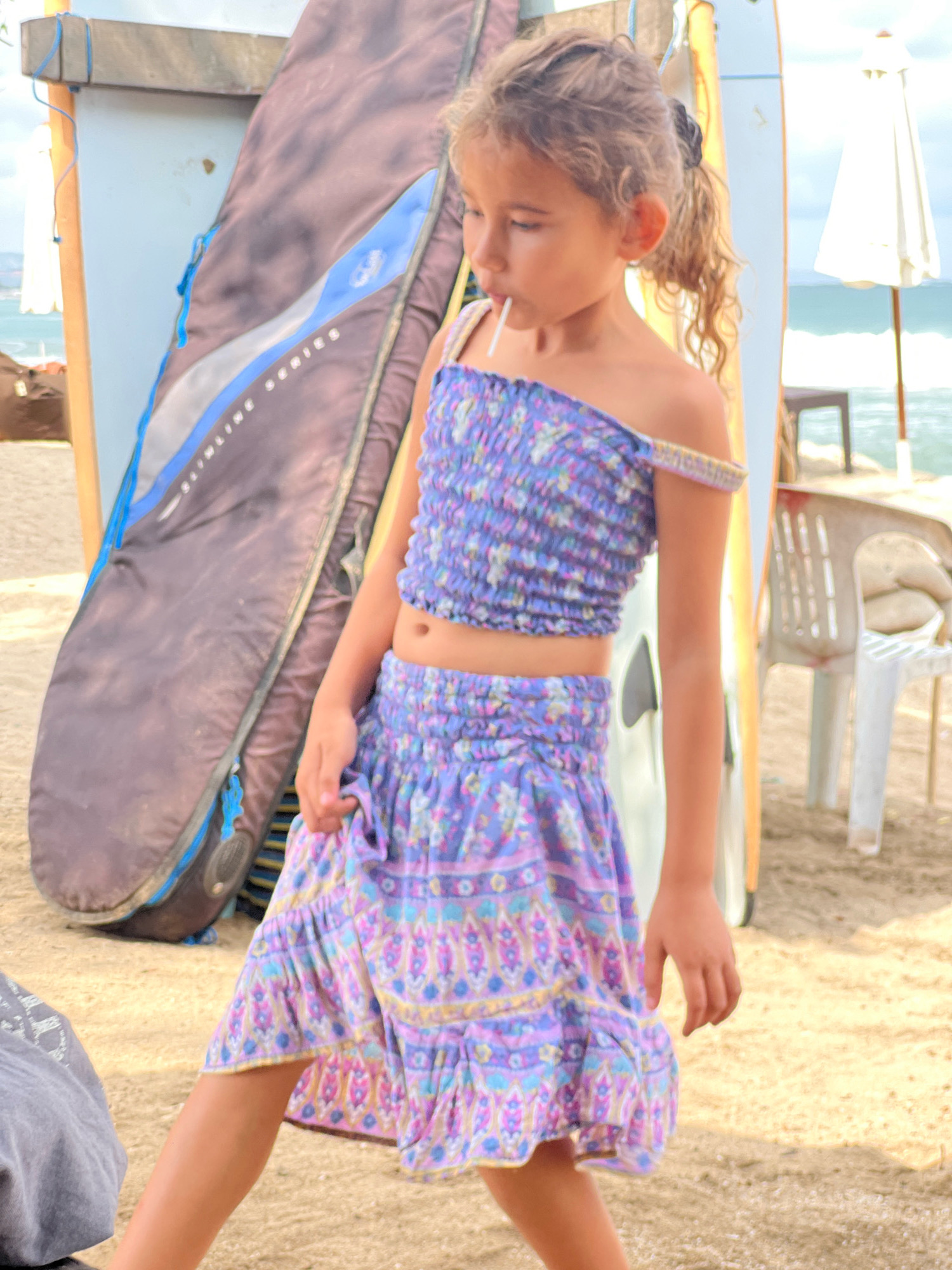 A young girl with curly brown hair pulled back in a ponytail, wearing an off-the-shoulder purple floral print crop top and a matching tiered skirt, stands on a sandy beach in Kuta, Indonesia. She has a white lollipop in her mouth and is looking to her left. Behind her, a dark blue surfboard bag leans against a wooden structure. To her right, a pale yellow surfboard and a white beach umbrella are visible, along with a white plastic chair. The ocean with waves is in the background. A partially visible gray shirt is in the lower left corner of the image.
