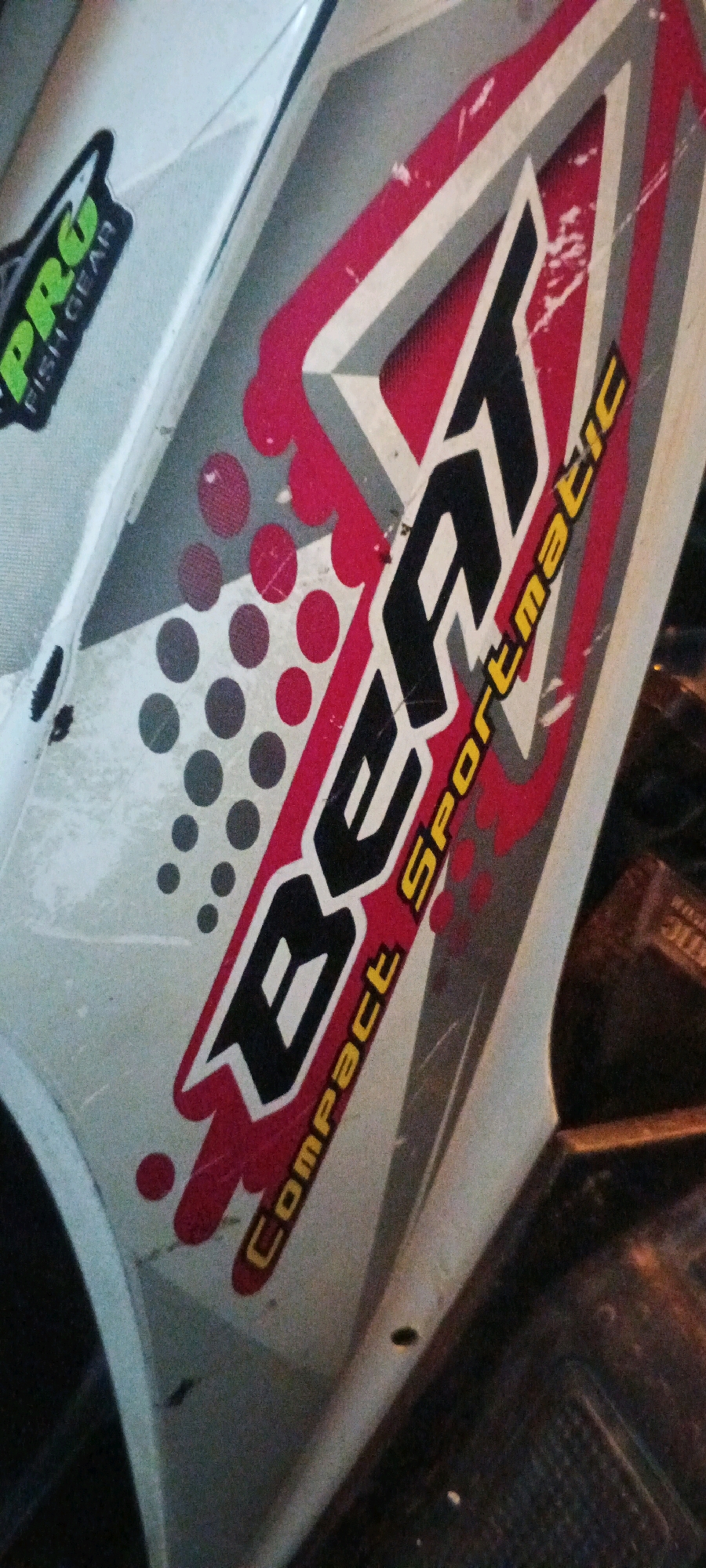 The image is a close-up shot of a white and red motor scooter decal. The decal features the word "BEAT" in large black letters with white outlining, followed by "Compact Sportmatic" in yellow lettering. A smaller decal on the left side of the image reads "PRO FISH GEAR" in green and black lettering. The background is dark and out of focus. The image is taken at a slight angle, showing the curved surface of the scooter. The overall impression is of a close-up of a vehicle's branding. There are no people or environmental details visible that would indicate a specific location in Kesiman, Indonesia.