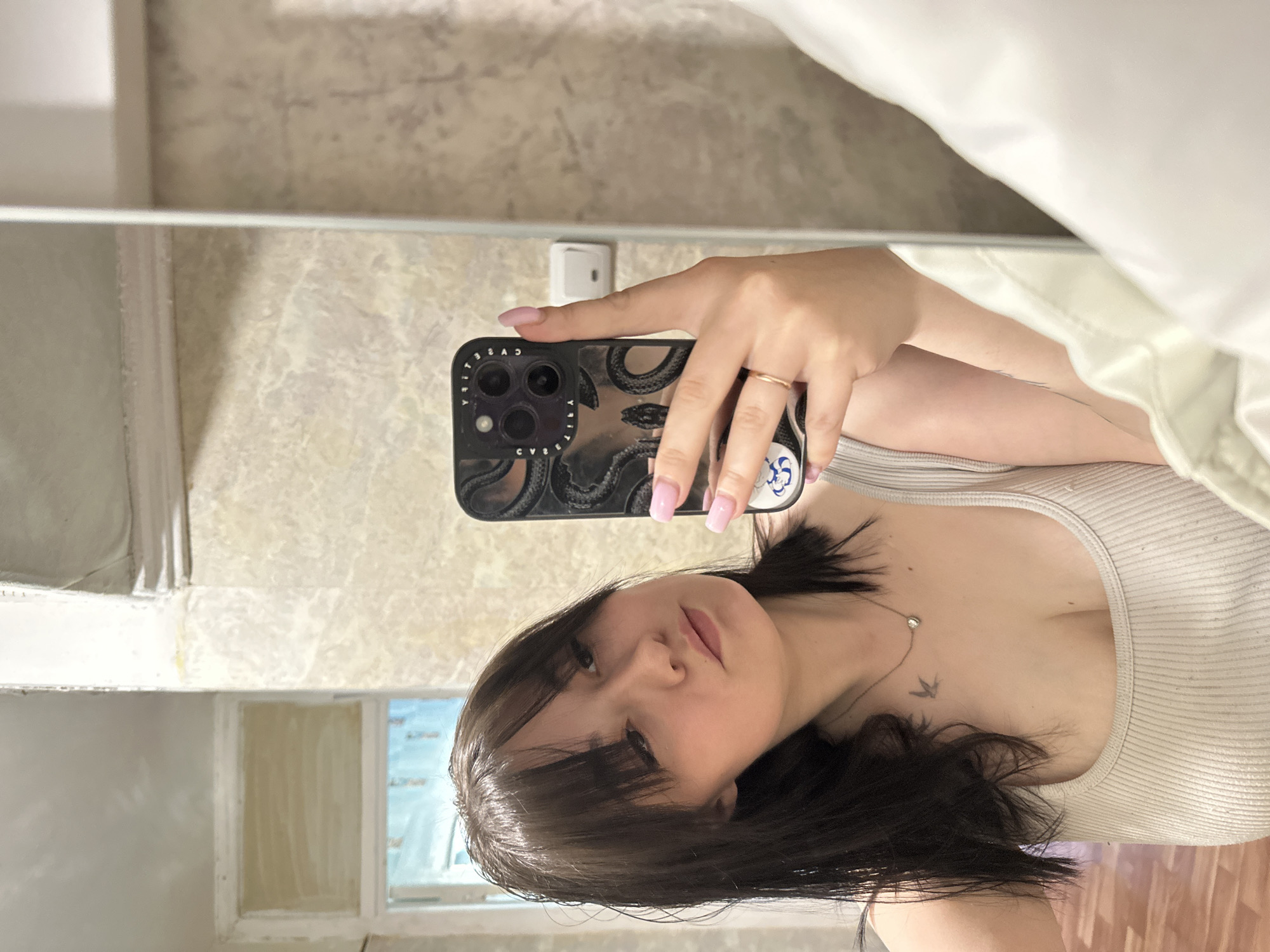 The image is a selfie taken in a room. A woman with dark hair, wearing a light-colored top, is holding a phone with a black case featuring a snake design. The phone is positioned to capture her face and upper body. A necklace with a small pendant and a small bird tattoo on her chest are visible. The background shows a mirror reflecting a room with light-colored walls and a window. The mirror frame is visible on the left side of the image. A light switch is seen on the wall behind the phone. The location is unspecified and cannot be determined from the image alone; therefore, specifying the location as Chaikovsky, Russia is not possible based on the visual evidence.
