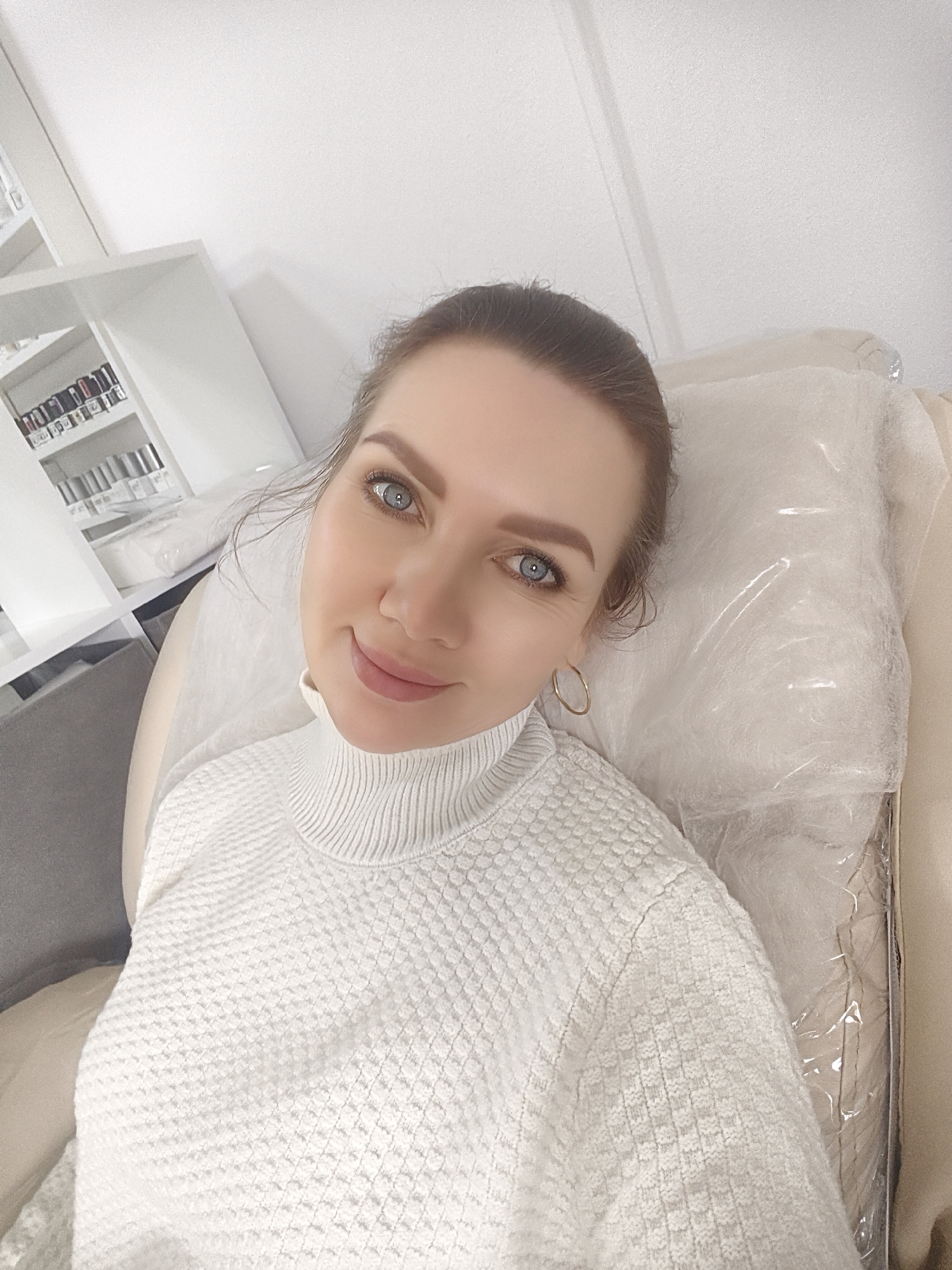 A woman with fair skin and blue eyes, wearing a white textured turtleneck sweater and gold hoop earrings, is pictured from a high angle. Her hair is pulled back. She is reclining in a padded chair covered with clear plastic. Behind her to the left is a white shelving unit containing small bottles or containers. The location appears to be an indoor space, possibly a beauty salon or clinic in an unidentified city and state.