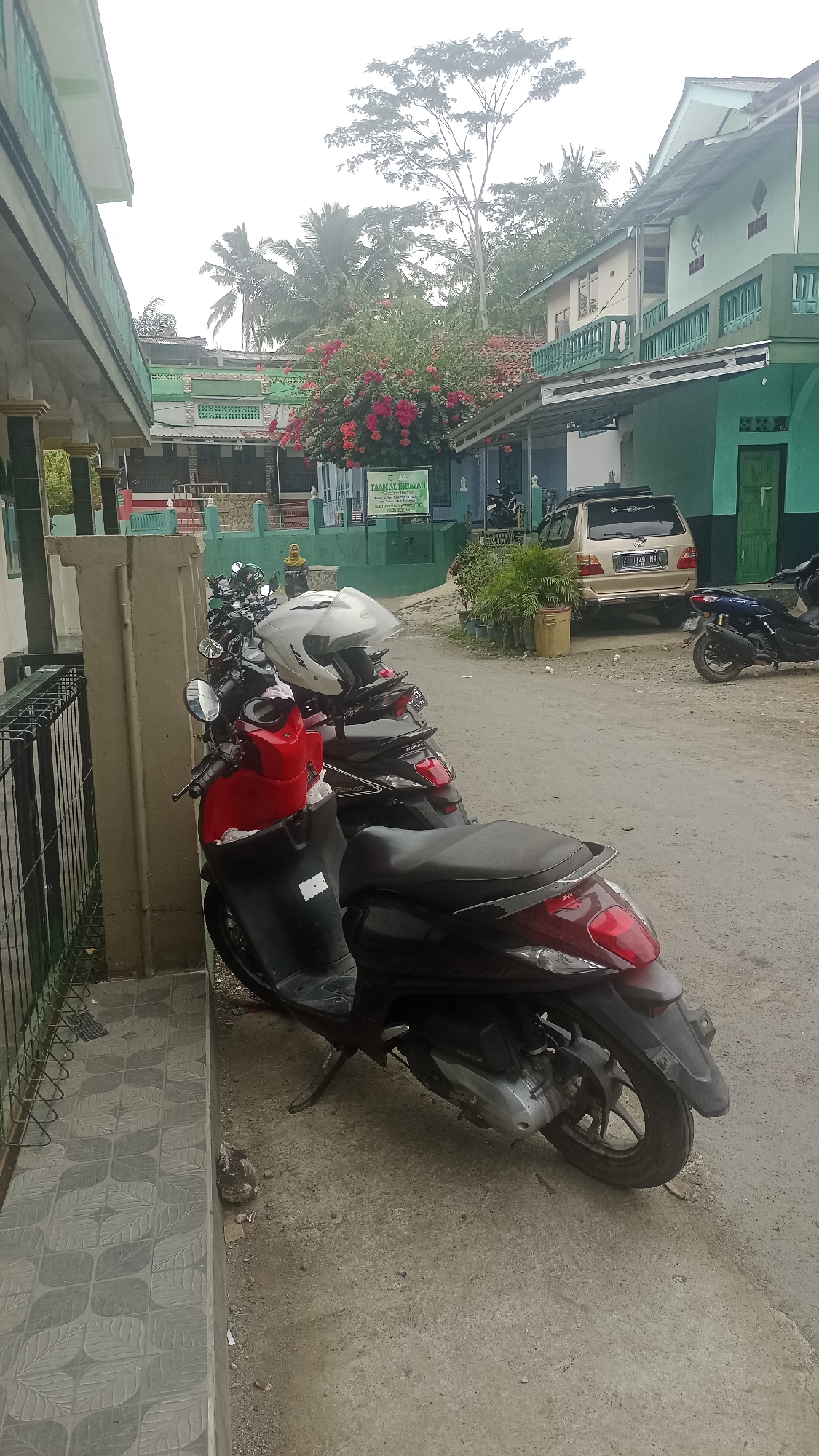 This is a photograph of an outdoor scene in Karyamandala, Indonesia. In the foreground, a row of motorcycles is parked along a walkway. The closest motorcycle is red and black, with a white helmet resting on its seat. Behind it, several other motorcycles are visible, some dark-colored. To the left, a building with green trim and columns lines the walkway. In the background, more buildings with green and beige facades are visible, along with trees and vegetation. A gold minivan is parked on a dirt road to the right of the motorcycles. A sign with Indonesian text is posted on a post in the middle ground, and a person wearing a yellow hijab is visible walking in the distance. The sky is overcast.