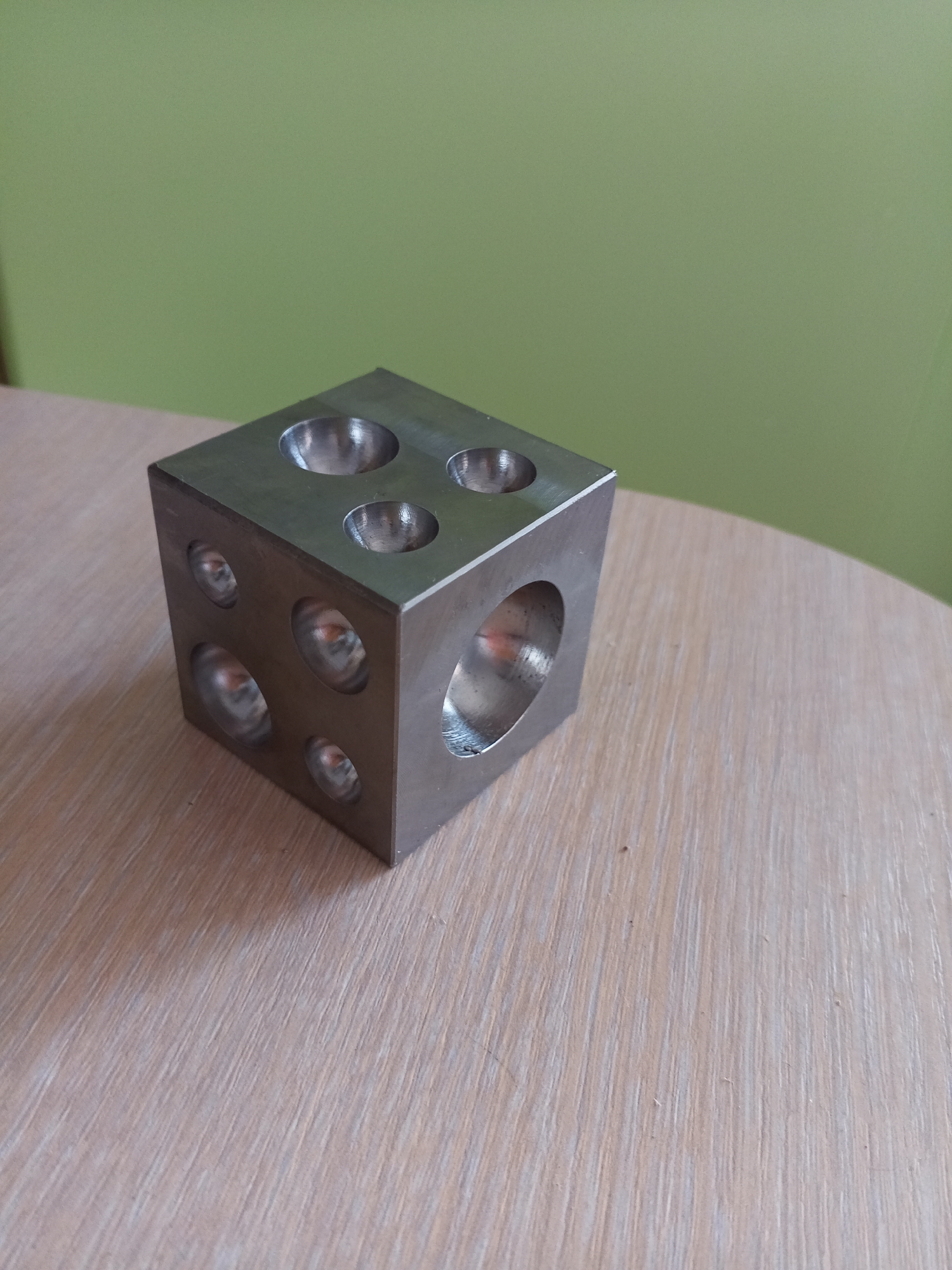 A metal cube with hemispherical indentations on multiple faces sits on a wooden surface against a light green background. The cube has four indentations visible on the top face and three visible on the left face. One large, circular opening is visible on the front right face. The wooden surface exhibits a light brown color with a visible wood grain pattern. The background is a solid, light green color. There are no people or animals present, and no discernible actions or interactions are taking place. The scene appears to be indoors, possibly in a workshop or studio. The specific location in the city of Vilyuysk, Russia, cannot be determined from the visual information provided.