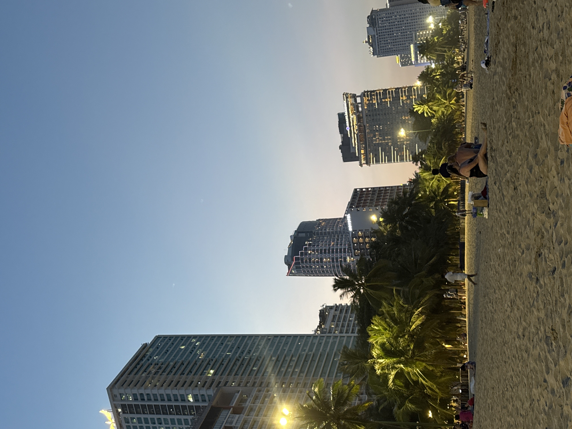This image captures a beachfront scene in Nha Trang Ward, Vietnam, during twilight or early evening. The sky exhibits a gradient, transitioning from a deep blue at the zenith to a lighter, paler blue with a subtle orange tint near the horizon. A faint celestial body is discernible in the upper right quadrant of the sky.
The background is dominated by a skyline of multiple tall, modern buildings, primarily hotels, which line the coast. Many of these structures display numerous illuminated windows and exterior lighting. On the far left, a prominent building features a distinctive, illuminated yellow, wing-like structure on its rooftop. Towards the center-right, signage for "Novotel" is visible on one building, and slightly to its left, another building bears the "Hyatt Regency" logo.
Between the buildings and the sandy expanse, a dense line of palm trees stretches across the midground. Several of these trees are illuminated from below or behind by artificial lights, casting bright green and yellow hues.
The foreground and midground consist of a wide, light-brown sandy beach, showing evidence of footprints and slight undulations in the sand. Artificial illumination from streetlights positioned within the palm grove and along the beachfront casts yellow light onto sections of the sand and foliage.
Numerous individuals and small groups are present on the beach. In the immediate foreground, slightly to the right of the center, a couple is seated on the sand; one individual wears a dark-colored swimsuit. To their left, various personal items, including what appears to be a cooler and bags, are placed on the sand. Further back on the beach, a person dressed in white is seen walking from left to right. Other individuals and small groups are scattered across the beach, some seated on the sand with towels or blankets, while others are standing or congregating near the tree line, extending towards the right side of the frame. On the far left of the beach, another small group is visible, including a person wearing a pink top.