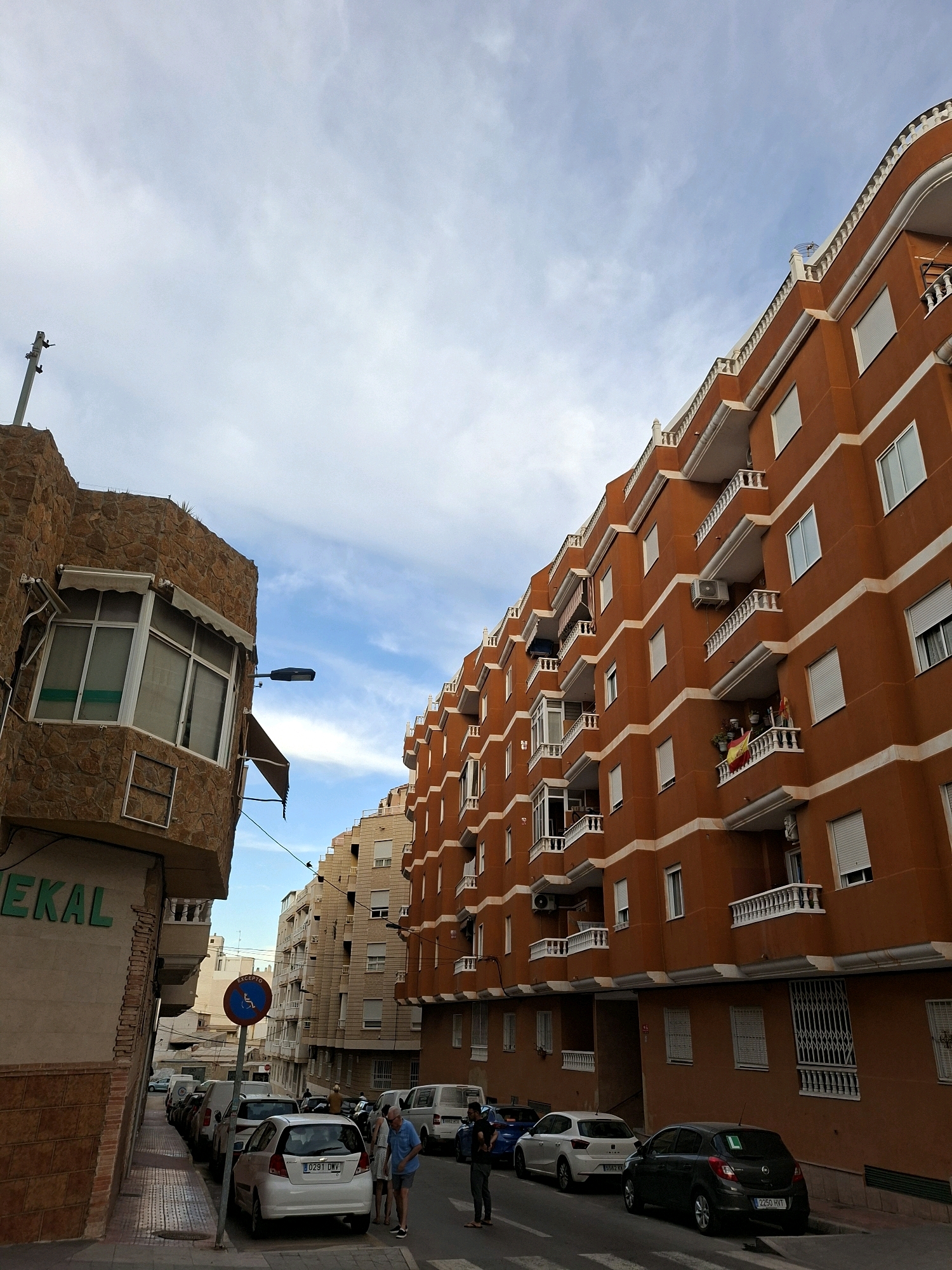 The image depicts a street scene in Torrevieja, Spain, with several buildings visible. On the left, a building with a textured stone facade features a prominent bay window with awnings. Below the window, green lettering spells out "EKAL". A street light is mounted on the side of this building.
The main focus of the image is a large, multi-story apartment building painted a vibrant orange. It has numerous balconies with white railings and decorative white trim along its edges. Several windows with white shutters are visible. On one of the balconies, a Spanish flag is displayed. An air conditioning unit is mounted on the exterior wall of this building.
To the right of the orange building, another apartment building with a lighter, beige facade is partially visible. This building also appears to have multiple stories and balconies.
In the foreground, several cars are parked along the street. A white hatchback with license plate "0291 DMV" is prominently positioned. A dark-colored hatchback with license plate "2250 HVT" is parked behind it. Further down the street, other vehicles, including vans and cars, are lined up.
A blue and white circular traffic sign with a red border is visible on a pole, indicating a prohibition. Two individuals are standing near the white hatchback, and another person is visible further down the street.
The sky above is partly cloudy with patches of blue sky visible. The scene appears to be an urban environment, likely a residential area.