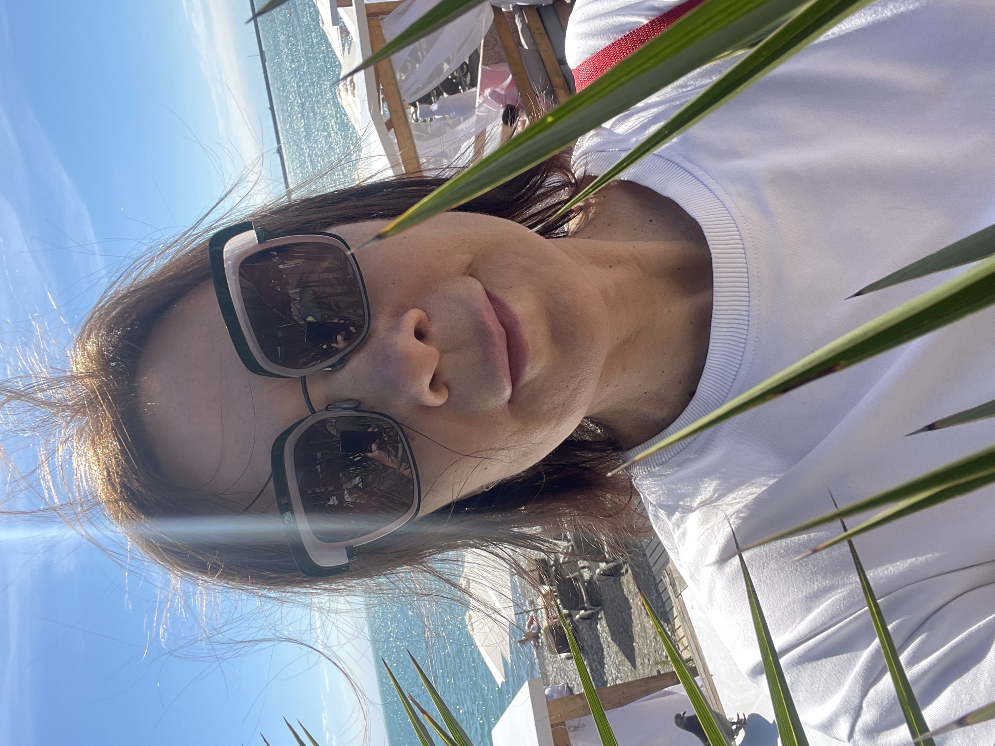 A close-up, portrait-oriented image features a woman posing outdoors on a sunny day, likely in a coastal area like Sochi, Russia. She has dark brown hair, parted on the side, and is wearing stylish oversized sunglasses with dark lenses that reflect an indoor or shaded area. A slight, contented smile is visible on her lips, and her complexion is warm-toned. She is dressed in a plain white crew-neck sweatshirt or t-shirt, with a hint of a red strap from a bag or garment visible on her right shoulder.
In the immediate foreground, several vibrant green palm leaves partially frame the shot, adding a tropical feel. The background is dominated by a bright blue sky with a few faint clouds and the sparkling blue waters of the sea, suggesting a beautiful beach or waterfront setting. Further back, white beach chairs or sun loungers under white coverings can be seen, indicating a resort or beach club environment. The strong daylight creates bright highlights on her hair and the water, confirming it is daytime. The overall mood conveyed is relaxed and pleasant. There is no visible text in the image.