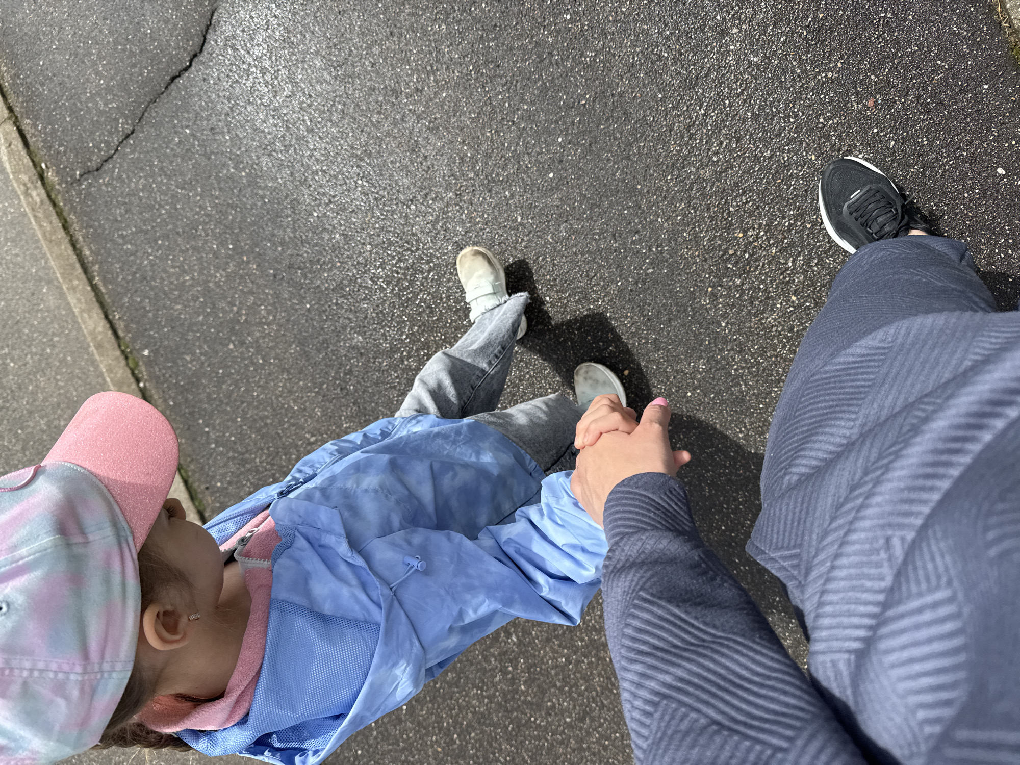 A child wearing a pink and tie-dye baseball cap, light-blue jacket over a pink hoodie, and light-wash jeans with white shoes walks hand-in-hand with an adult on a paved surface. The adult wears black and white sneakers and a blue textured pant and shirt. The surface appears to be asphalt with a cement curb to the left. The location of the scene is not mentioned in the prompt, so I cannot include the city of Shevlyakovo, Russia.