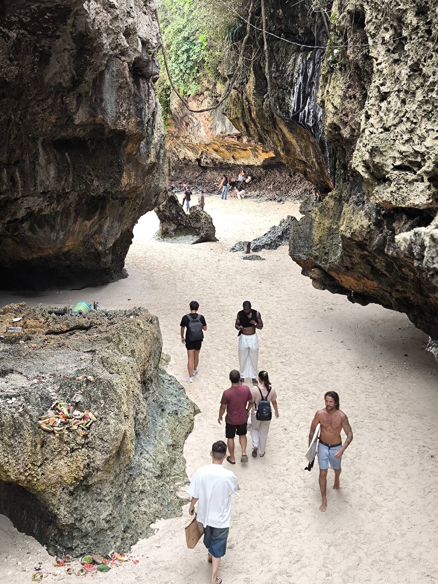 The image shows a sandy beach framed by rocky cliffs, likely a cove. The location is Uluwatu, Indonesia, based on the presence of cultural offerings (Canang Sari) and the general environment. A group of people are walking on the sand, primarily in the lower half of the image. In the foreground, a man with a surfboard is walking toward the viewer. Behind him, a small group of three people walks in the same direction. Farther away, two more people are walking away from the viewer. In the background, another group of people are congregating on the sand near the cliffs. The cliffs are made of porous rock, with some vegetation visible on top. On the left side, there is a small rock formation with a Balinese offering on top. The overall environment appears tropical.