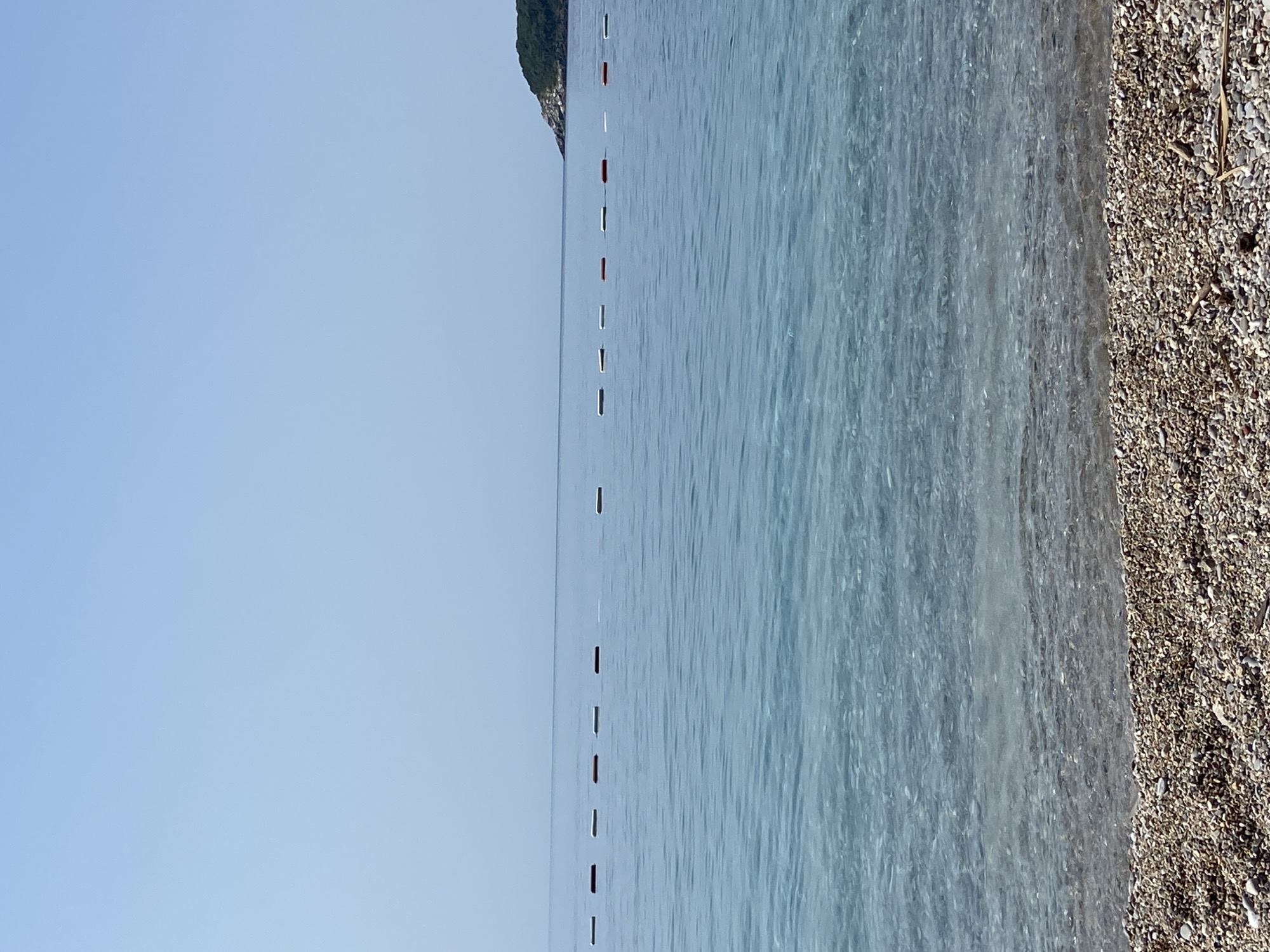The image shows a tranquil scene at the beach in Prijevor, Montenegro. The foreground consists of a pebble beach, with the pebbles varying in size and color, interspersed with small pieces of driftwood. The sea occupies the middle ground, its water a clear, shimmering turquoise, allowing the pebble seabed to be visible in the shallower areas. A line of buoys, alternating between white and red, marks a boundary in the water. In the background, the calm sea meets a pale blue sky at the horizon. To the far right, a lush green, tree-covered headland juts out into the sea, its rocky base partially visible. The overall atmosphere is serene and peaceful, with no people or animals present in the frame.
