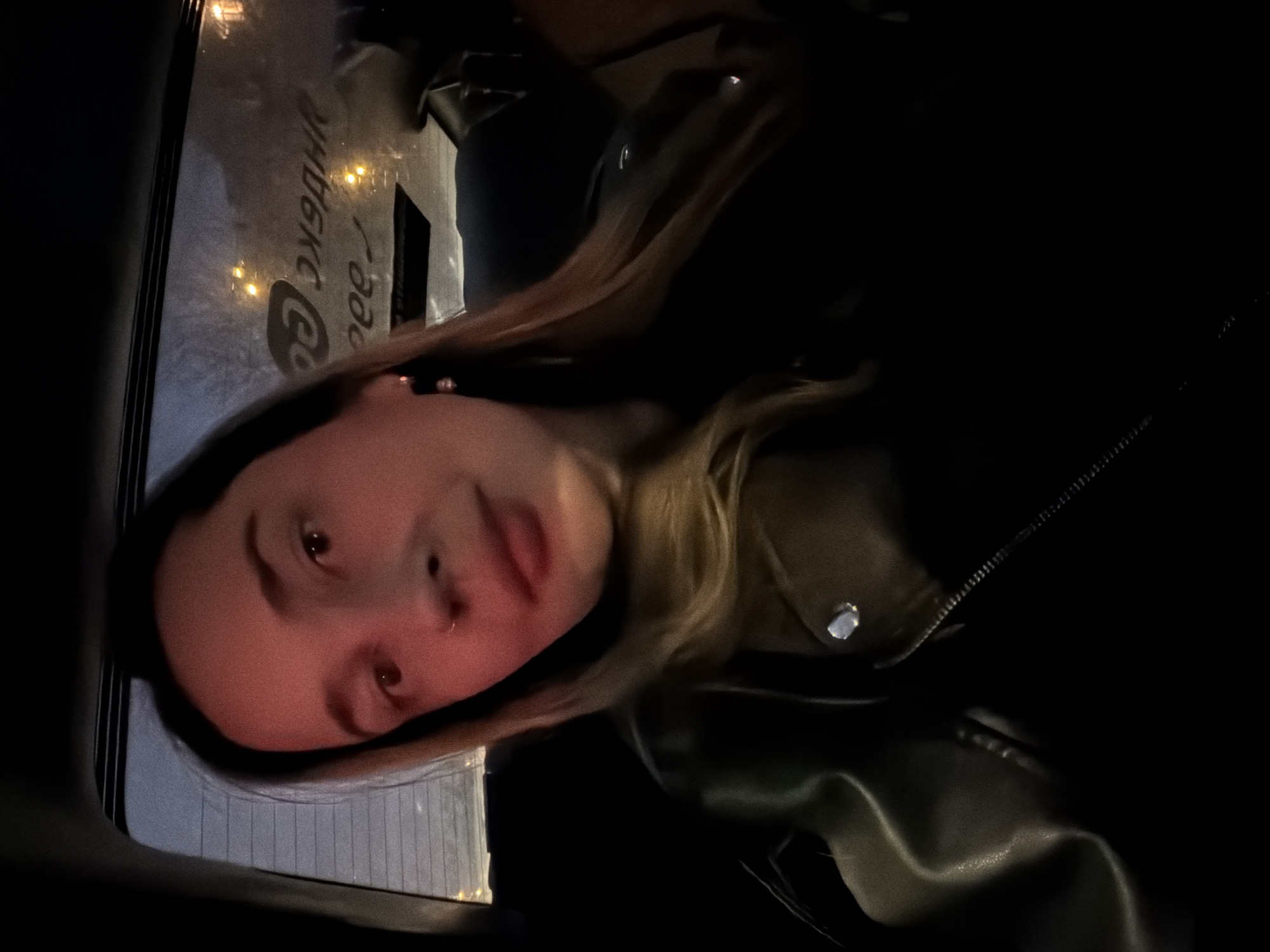 A young woman with brown hair is visible in the low-light image. She has a nose piercing and is wearing a dark leather jacket. The image appears to be taken in a car, and a window with Russian text ( likely reading "Yandex Go") can be seen, suggesting the location is in Moscow, Russia. The picture may be a self-portrait.