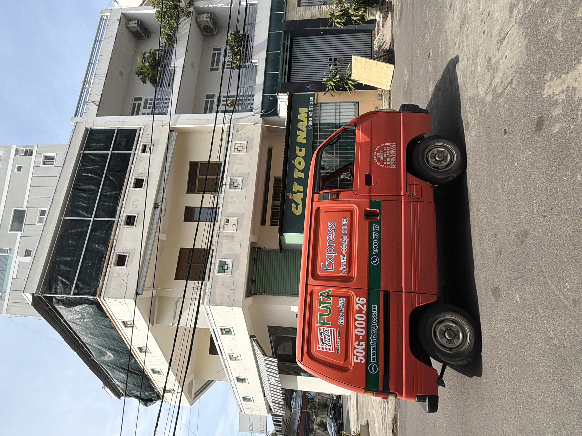 A bright red FUTA Express van is parked on an asphalt street in front of a multi-story building. The van has the FUTA Express logo and other text and graphics on its side, including "50G-000.26" and "www.futaexpress.vn". The building behind the van appears to be a commercial establishment, with storefronts and windows. A sign above one of the storefronts reads "CẮT TÓC NAM" which translates to "Men's Haircut". The sky is blue with some clouds. The scene is set in the city of Nha Trang, Vietnam, likely on a sunny day.
