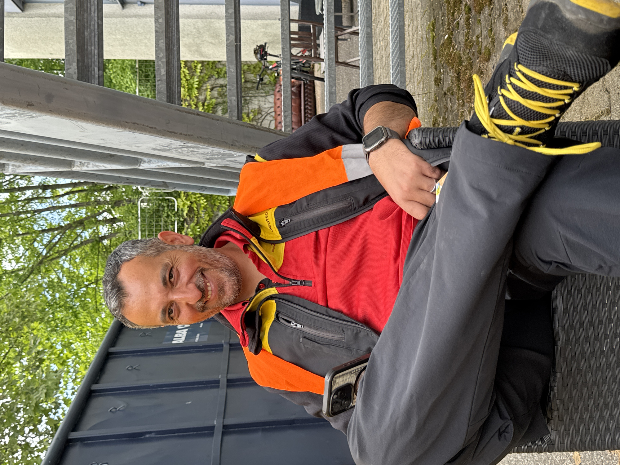 A man is sitting on a black wicker chair outdoors, possibly on a patio or terrace. He is wearing a red shirt with an open black and orange jacket, gray pants, and black shoes with yellow laces. He has a graying beard and is smiling. He is wearing a smartwatch on his left wrist and holding a gray cell phone in his left hand. The phone is partially visible, and there is a second cell phone in his jacket pocket.
Behind the man is a blue metal container with the word "ALBA" on it. Metal stairs and a railing are visible in the background. Further back, there are trees and a glimpse of a building. A bicycle is partly visible between the stairs. The ground appears to be concrete.
Given the presence of the ALBA container, the location is very likely in Bietigheim-Bissingen, Germany, as ALBA is a well-known German recycling company.