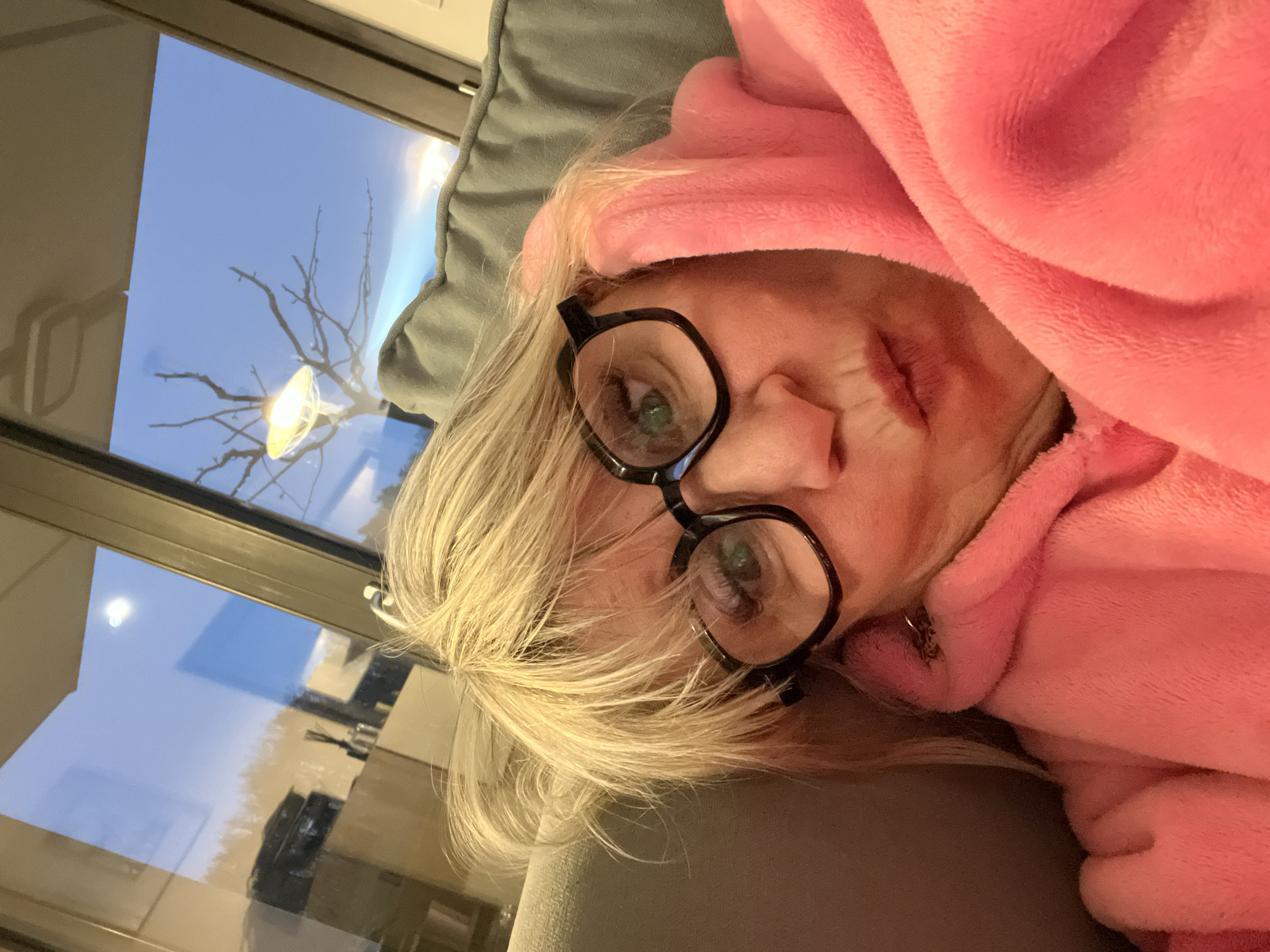A woman with blond hair, wearing black-framed glasses and a pink hooded robe, is lying on a gray surface. Reflected in the window is a tree branch and a street light. The location is Chambœuf, France.