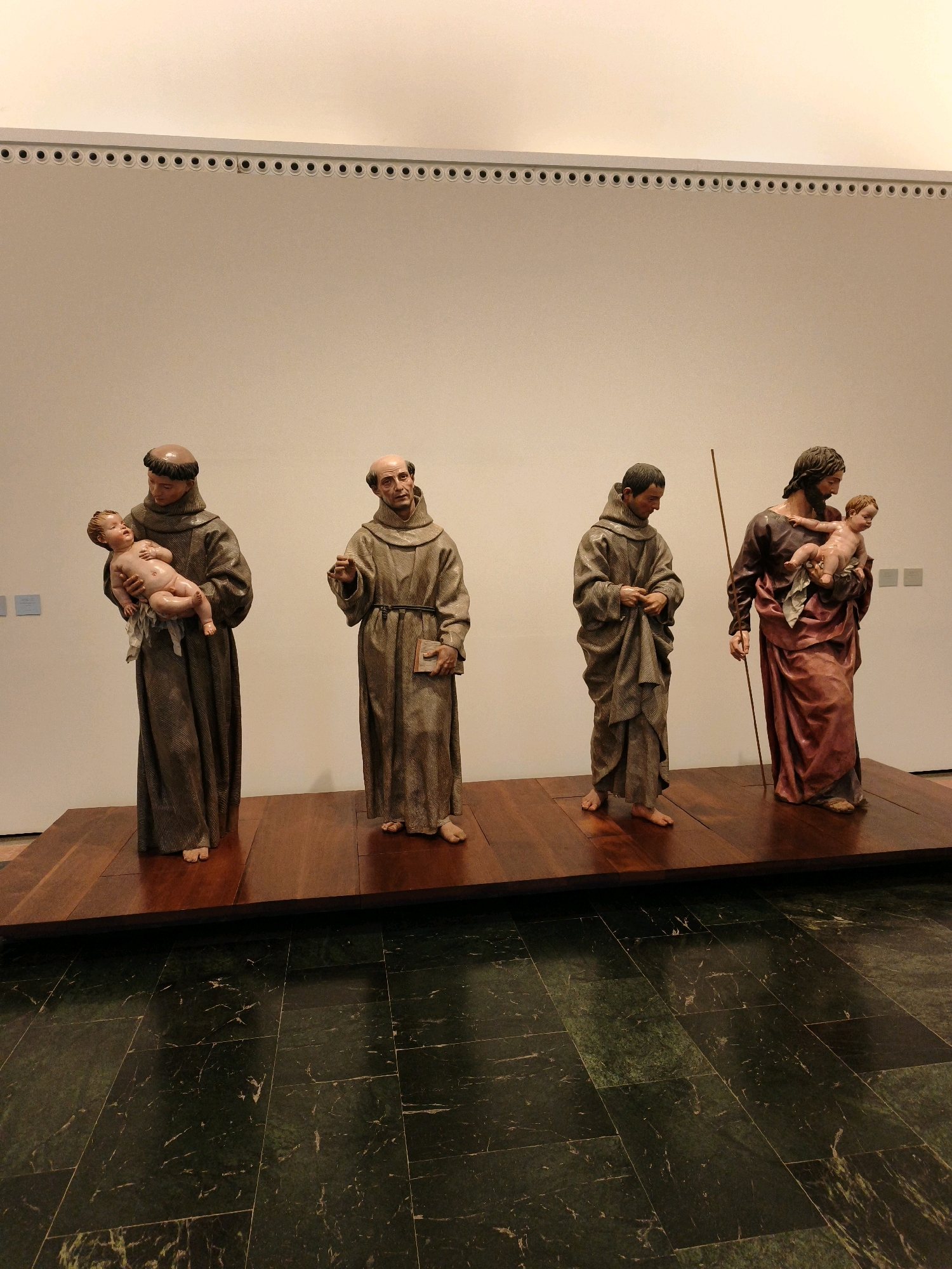 Four wooden statues of religious figures are displayed in a museum. The statues are arranged in a line from left to right. The first statue depicts a male figure in a brown robe holding a baby. The second statue shows a bald male figure in a brown robe holding a book. The third statue portrays a male figure in a brown robe. The fourth statue features a male figure with dark hair, wearing a red robe, holding a baby, and carrying a staff. The statues are standing on a wooden platform. The background is a plain white wall with a decorative border along the top. The floor is a dark, polished stone with a marbled pattern. This scene is located in the Museo de Bellas Artes de Granada, located in the Palacio de Carlos V in Granada, Spain.