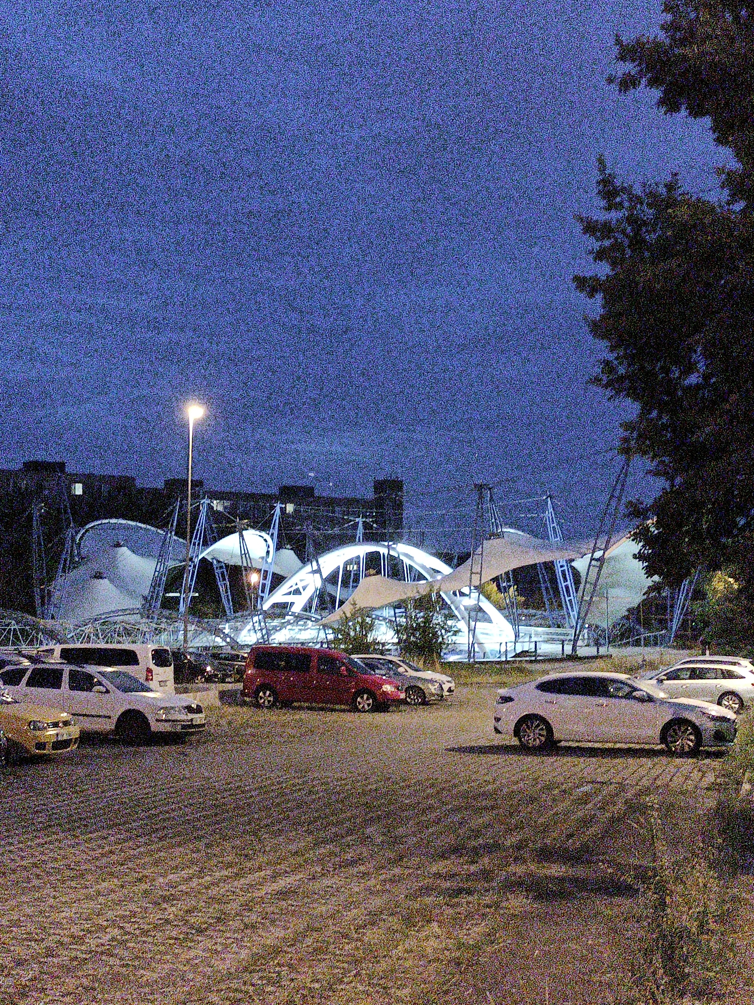 A nighttime scene captures a parking lot in front of a modern, illuminated architectural structure with a distinctive white, tent-like roof supported by metal poles and a bright street lamp. Several cars are parked in the lot, including a yellow Volkswagen, a white van, a red minivan, and a white sedan in the foreground. The background shows buildings, and trees partially obscure the view. The overall atmosphere is dimly lit, with the artificial lighting from the structure and street lamp dominating the scene. The image is grainy, suggesting low light conditions or a low-resolution capture.