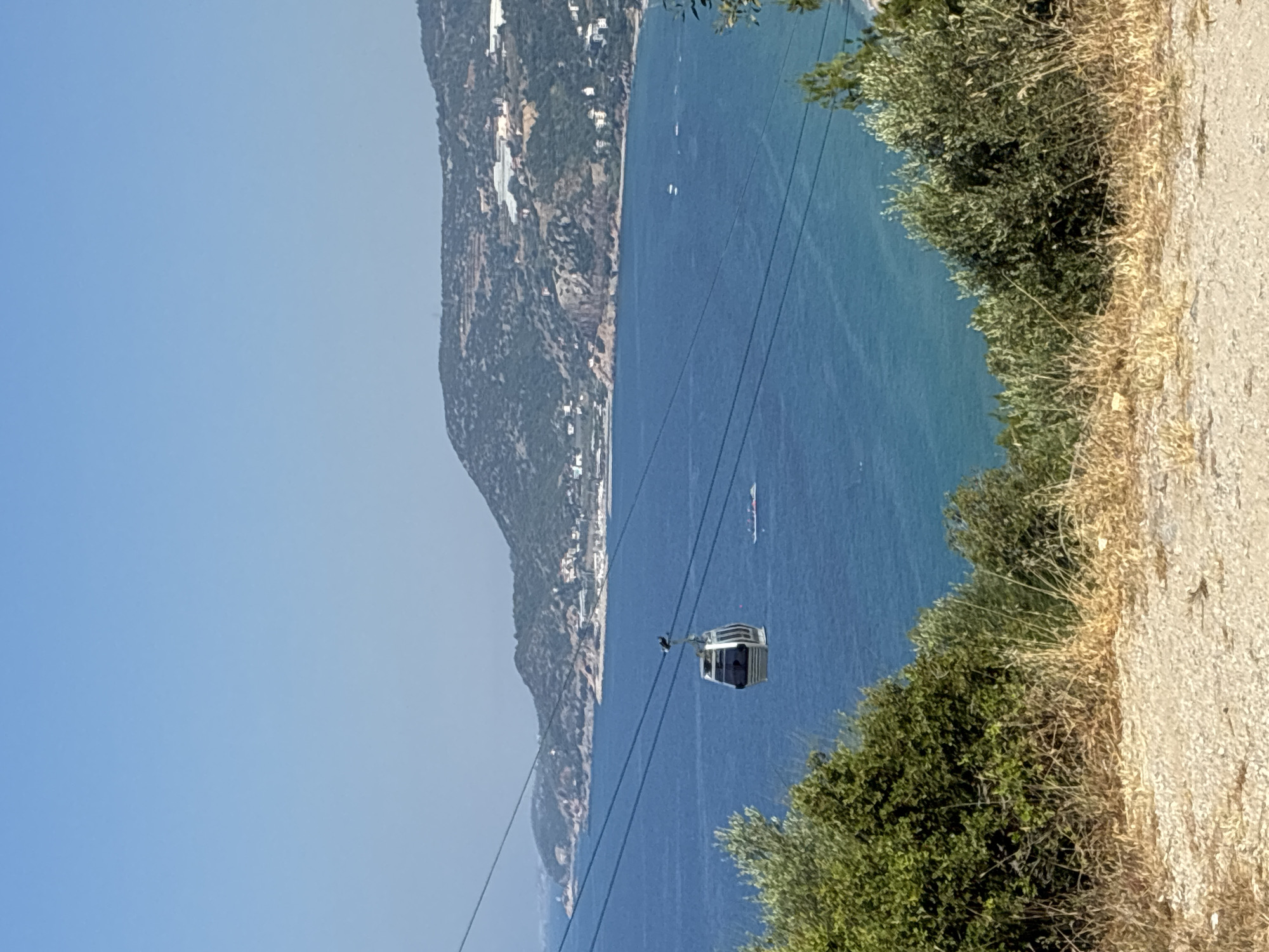 In Alanya, Turkey, a cable car traverses the clear blue sky above the Aegean Sea. The cable car, a modern, silver gondola, is suspended from thick cables that stretch across the frame, connecting to unseen points on the distant, verdant coastline. Below the cable car, the sea is a vibrant blue, dotted with a few small boats. The coastline itself is a rugged landscape of green hillsides, dotted with buildings and possibly greenhouses, characteristic of the region. In the foreground, the terrain is dry and scrubby, with patches of green bushes and dry grasses leading down towards the sea. The sky above is a cloudless, bright blue, indicating a sunny day.