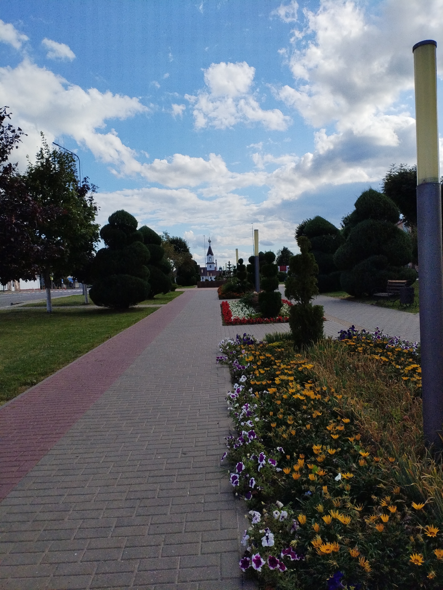 The image depicts a paved pedestrian walkway in Bialynichy, Belarus. The walkway is constructed from grey rectangular pavers with a section of red pavers forming a border. It is lined on one side with a neatly manicured lawn and trees, and on the other side with flower beds. The flower beds contain a variety of colorful flowers, including yellow daisies and purple and white petunias. In the distance, a white building with a steeple, likely a church, is visible. The sky is blue with scattered white clouds. A lamppost stands prominently on the right side of the walkway. The scene appears to be a public park or a landscaped area within the city. There are no people visible in the image.