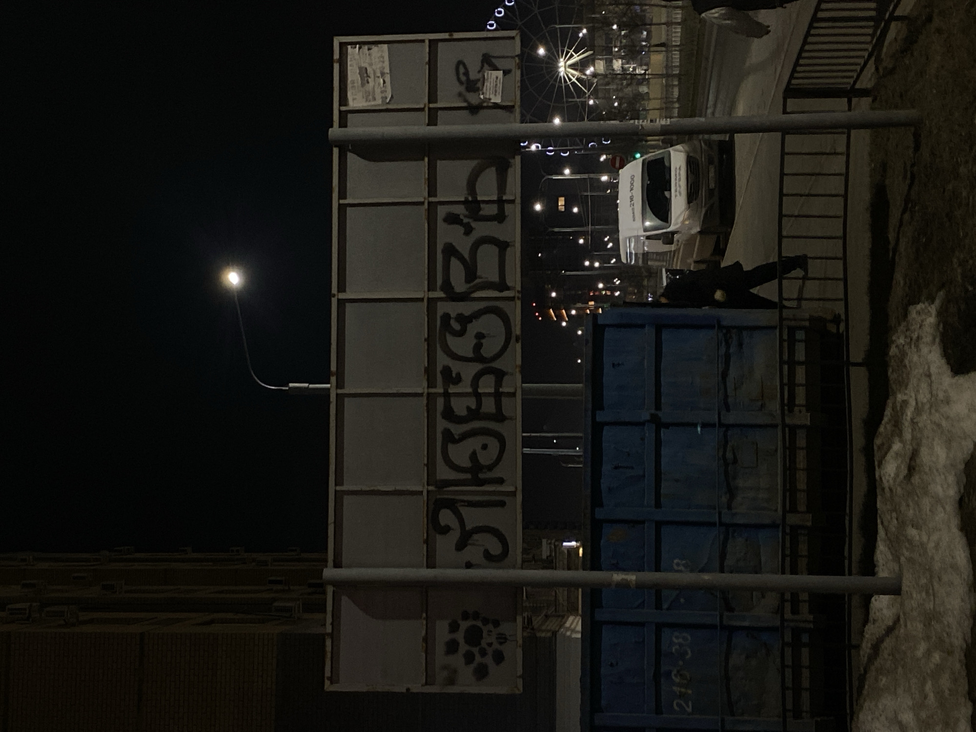 Night shot from Kazan, Russia. A street light illuminates an urban scene with a large sign covered in graffiti. A utility truck is parked on the street. A person dressed in dark clothing is walking nearby. A blue metal structure with the marking "216-38" is near the sign. A Ferris wheel is visible in the background. Buildings with multiple windows are in the distance. Snow is on the ground.
