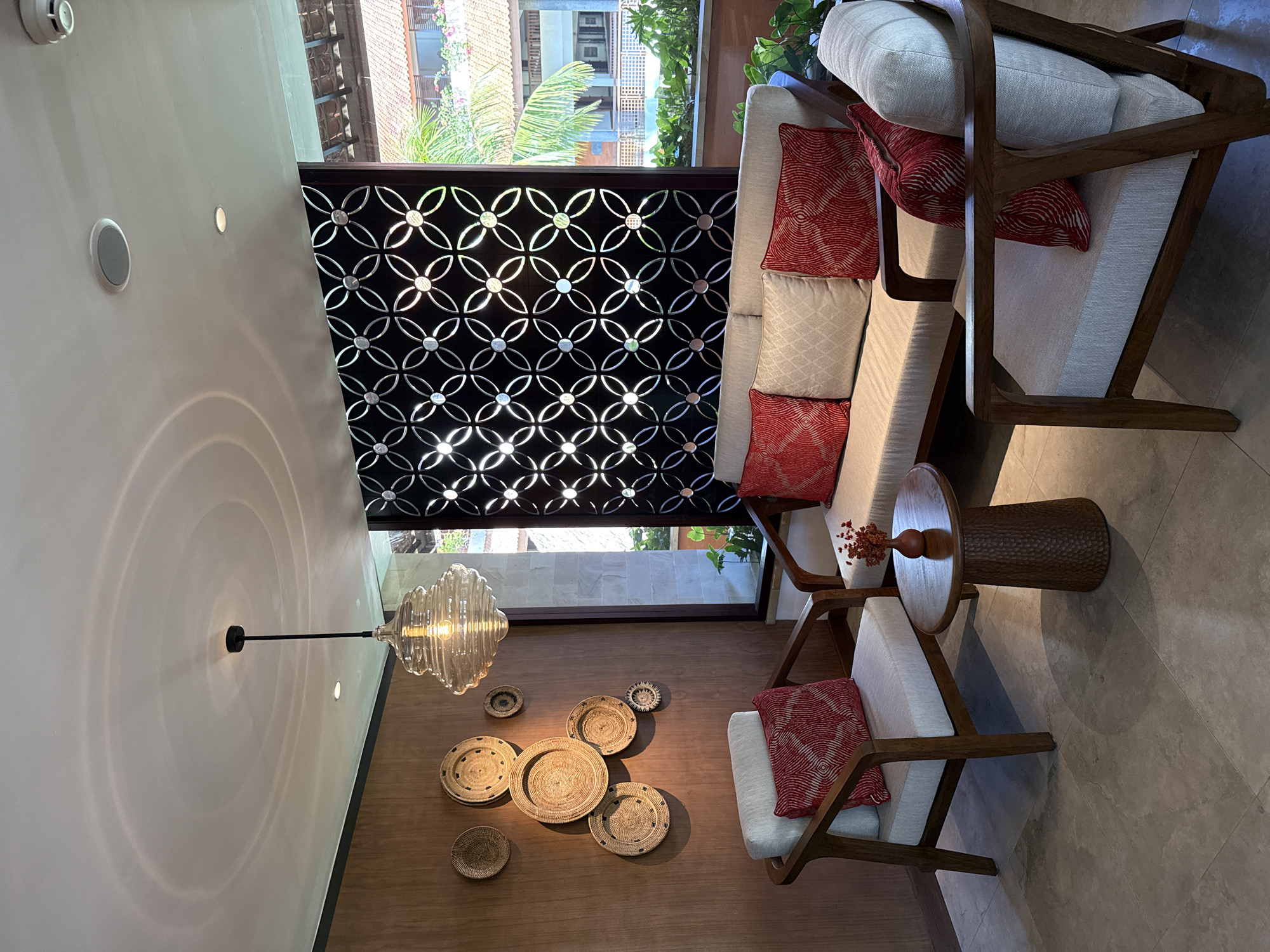 The image shows the interior of a space in Kuta Selatan, Indonesia. The space features two light-colored sofas with wooden frames and red patterned throw pillows. There is a wooden side table between the sofas. A wall features a geometric pattern screen. On the adjacent wall, decorative woven baskets are displayed, and a glass chandelier hangs overhead, casting concentric light patterns on the wall. A glimpse of the outside environment reveals tropical greenery and building structures, typical of the area.