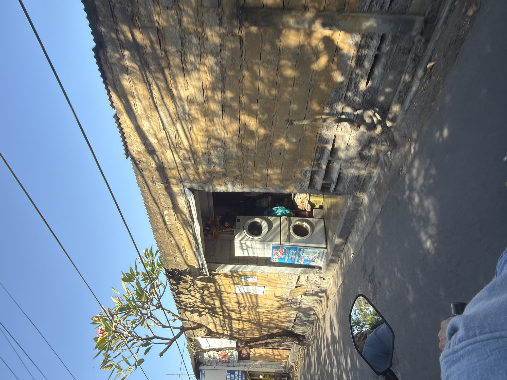 An exterior, eye-level, medium shot captures a roadside laundry service in Denpasar, Indonesia. The scene is dominated by a rough, yellow-hued stucco wall, with a corrugated metal roofline visible at the top. The wall is textured with horizontal lines, suggestive of stacked bricks or logs, and dappled with the shadows of tree branches.
In the center of the image, an open doorway reveals the interior of the laundry. Two stacked white washing machines are positioned just outside the entrance. A blue and white sign with text is propped against the wall near the washing machines. Inside the doorway, obscured by shadows, are piles of laundry or fabric.
To the left of the laundry shop, a narrow building with a sign is visible, indicating a business with the words "NARUNG" and "NASI CAMPUR" in Indonesian. A tree with green leaves and a few pink flowers stands between this building and the laundry shop, its branches casting intricate shadows on the wall. Power lines crisscross the clear blue sky above.
In the foreground, the handlebar and mirror of a motorcycle are visible, along with a hand wearing a blue garment. The mirror reflects the rider, also wearing blue, and a bright, sunny environment with trees and buildings. The motorcycle is on an asphalt road, with a curb and sidewalk to the right of the road. The overall impression is a snapshot of a typical street scene in a warm, sunny climate.