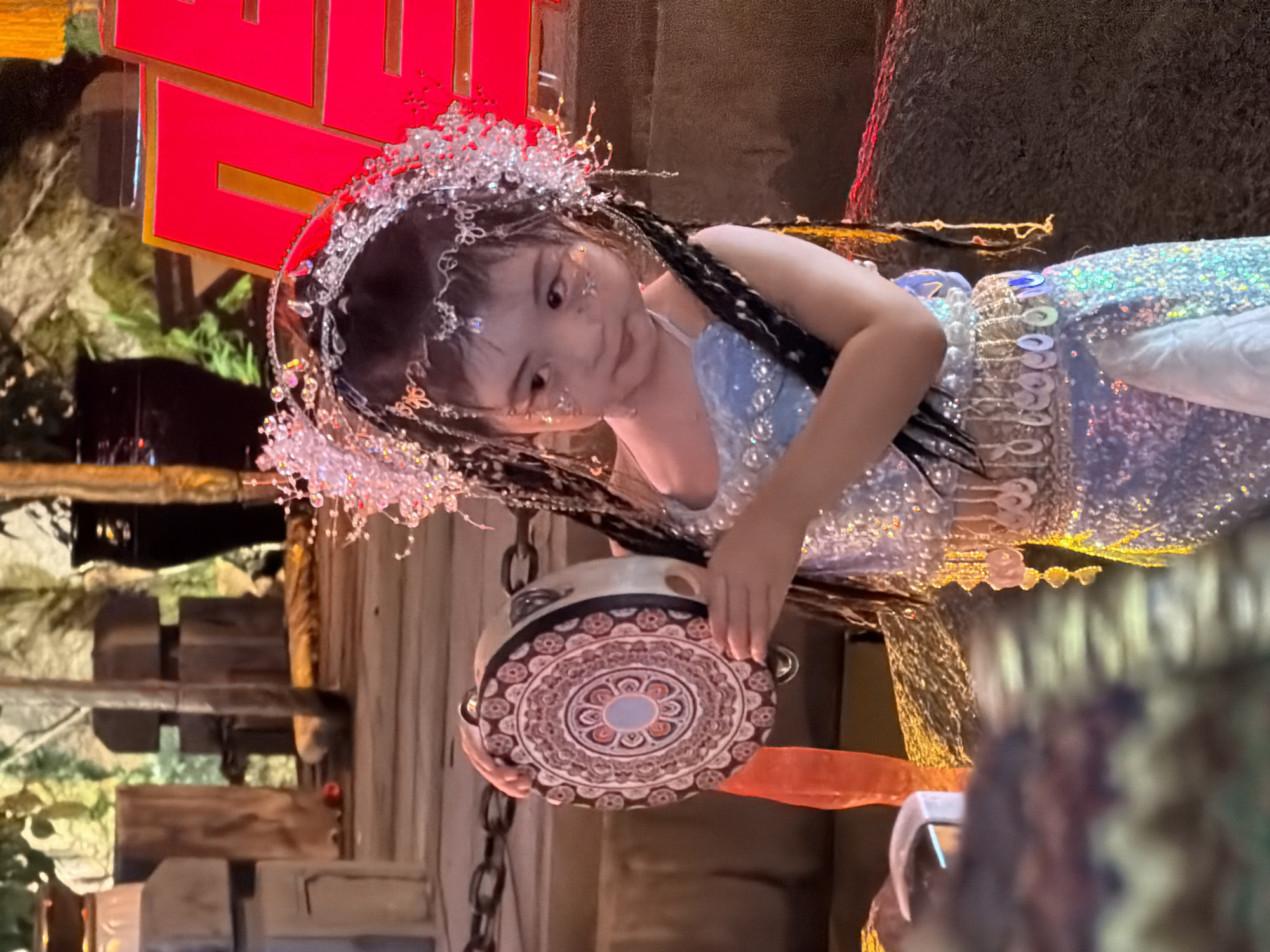 A young East Asian girl, adorned in a sparkly blue dress and an elaborate jeweled headdress, holds a patterned tambourine in her left hand. Her hair is styled in braids, and her face is decorated with glitter. She is standing outdoors, possibly in a market or festival setting in Yuzhong District, China, evidenced by the rustic wooden structures, chains, and large red signage in the background, which appears to spell out characters in Chinese. The lighting suggests it is either nighttime or dusk, with artificial lights illuminating the scene.