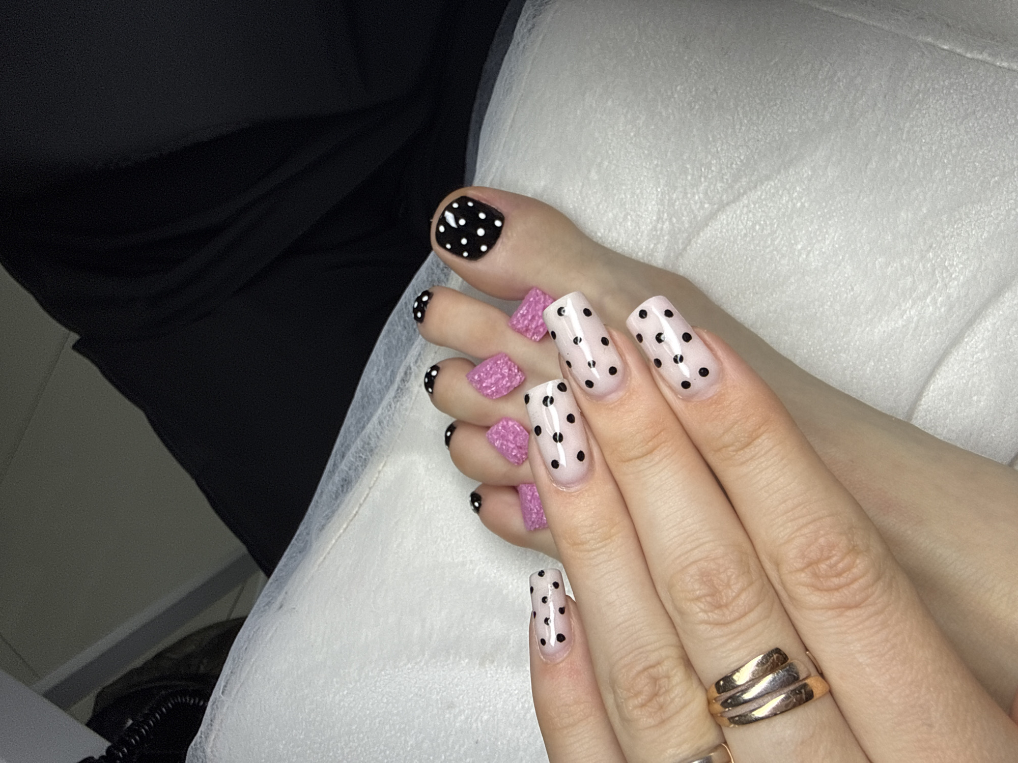 A person's hand is displaying a manicure with a pink base coat adorned with black polka dots. The fingernails are square-shaped. There are three gold rings on one of the fingers. The person's toes are also visible; the big toe has a black base coat with white polka dots, while the others have pink, textured decorations on them. The background is a white, textured surface, likely a cushion or towel. A dark garment is visible in the background, out of focus. There is no information about the location being Batumi, Georgia, or any indications of a city.