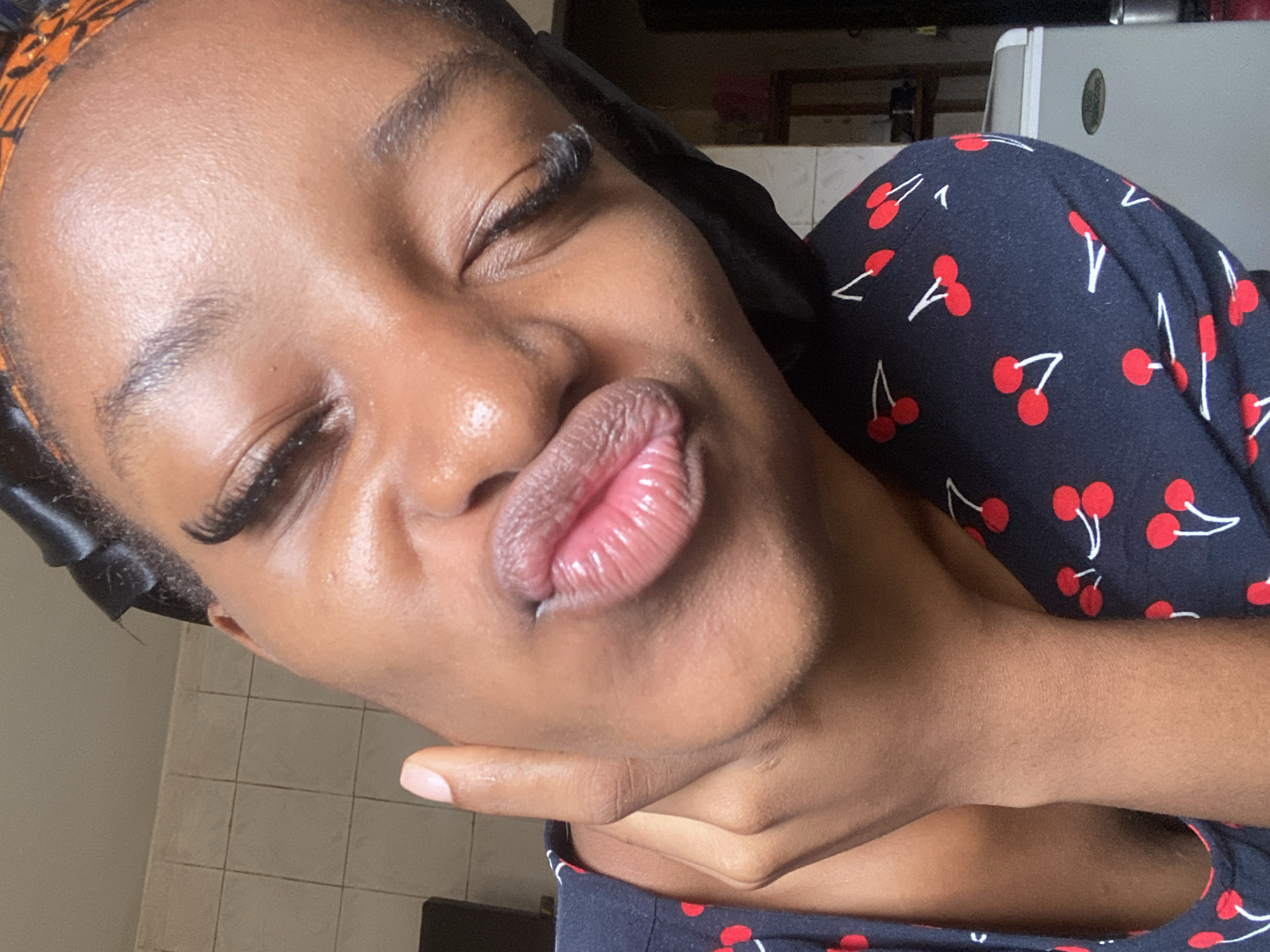 A dark-skinned individual is depicted in a close-up, angled shot, making a pouting 'kiss face' expression with glossy, pink-tinted lips. Her eyes are closed or mostly closed, featuring prominent, long false eyelashes. Her left hand is visible, with light pink nail polish on the thumb, supporting her chin and jawline. She wears a dark-colored garment patterned with red cherries and white stems. Her hair is covered by a dark, possibly satin, head wrap, with an additional patterned orange and brown fabric visible further back on her head. The background suggests an indoor setting. To the left, a light-colored tiled wall is visible. The right side of the background is blurred, showing indistinct objects, including a light-grey appliance (possibly a small refrigerator or cooler) and darker furniture or shelving. There are no visual indicators within the image to confirm the location as Jos, Nigeria.