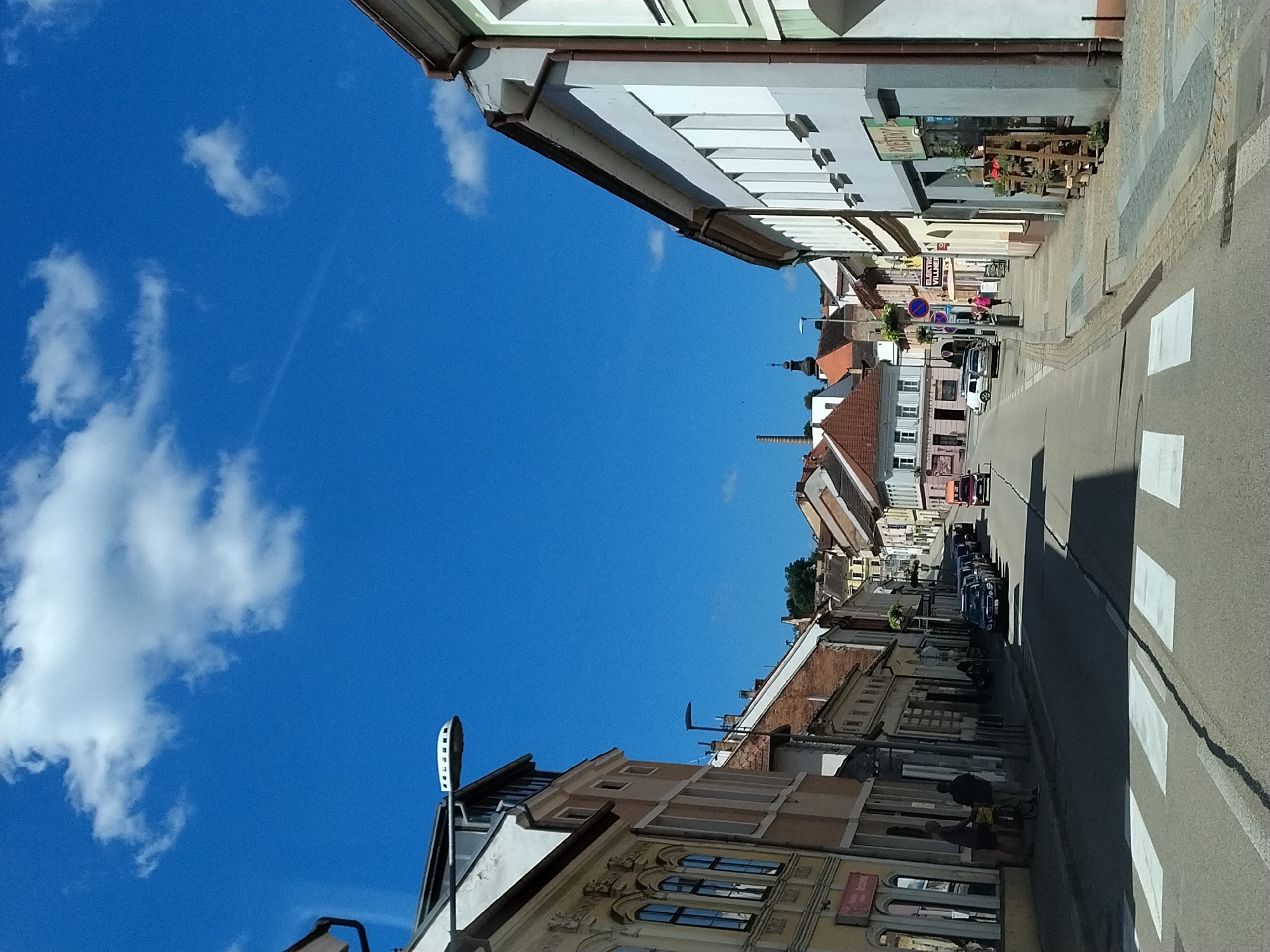 The image depicts a street scene in the city of Volyně, Czechia, under a bright blue sky with scattered white clouds. The perspective is from a low angle, looking down a cobblestone street lined with multi-story buildings. On the left, a pale yellow building with arched windows and ornate detailing is visible. To its right, a lighter colored building with a brown roof and several windows follows. The right side of the street features a building with a pale blue facade, white-framed windows, and a brown gutter system. In front of this building, a wooden display rack with plants is positioned on the sidewalk. Further down the street, several cars are parked along the side, including a black sedan, and an orange van is seen in the distance. Pedestrians are present, with two individuals standing on the sidewalk near the left-side buildings, one of whom appears to be interacting with a bicycle. Another pedestrian in a pink jacket is walking on the right-side sidewalk. A crosswalk is marked on the asphalt road in the foreground. In the background, the street curves upwards, revealing more buildings with red-tiled roofs and a distant church spire. A tall, brick industrial chimney is also visible in the background to the left. The overall impression is a sunny day in a European town.