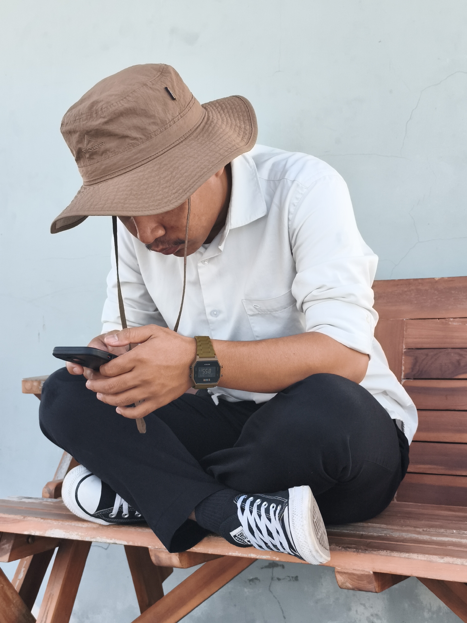In Semarang, Indonesia, a man sits cross-legged on a wooden bench. He wears a brown bucket hat, a white long-sleeved shirt, black pants, and black and white sneakers. A watch with a green band is visible on his left wrist. He is holding a black smartphone in his hands and appears to be looking at it. The background is a plain, light blue wall. The scene is lit by natural light, casting subtle shadows.