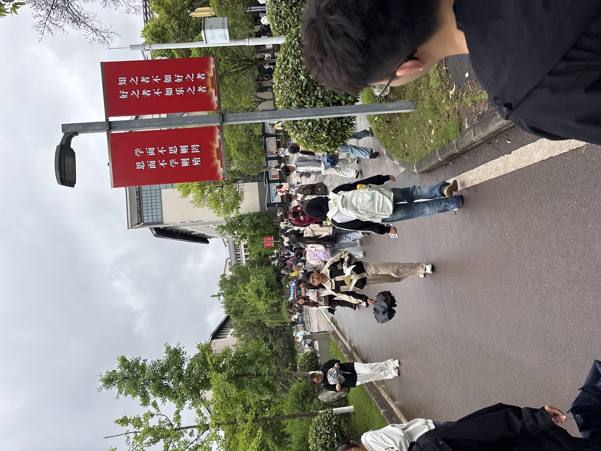 The image captures a scene in Jiangjin, China, with a crowd of people walking on a paved road or sidewalk lined with greenery. Red banners with white Chinese characters are prominently displayed on a pole. Trees and buildings are visible in the background under a cloudy sky. The crowd appears to be composed of students, perhaps on a school campus, dressed in casual clothing, some holding umbrellas. A person wearing glasses and a black jacket is partially visible in the foreground on the right side of the image.