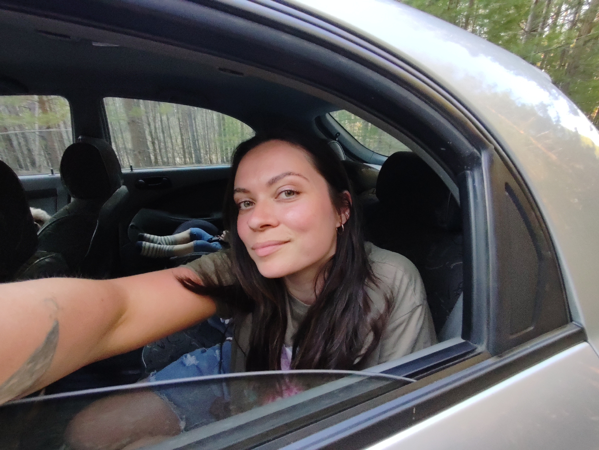 A medium shot depicts a young woman inside a car with the window partially lowered. She has long, dark hair and is wearing a light-colored shirt. She looks directly at the camera with a slight smile. The car's interior is visible, including the seats and other occupants, although they are partially obscured. The background shows a view of trees, suggesting the car is in a rural or wooded area. The location is not discernible from the image.