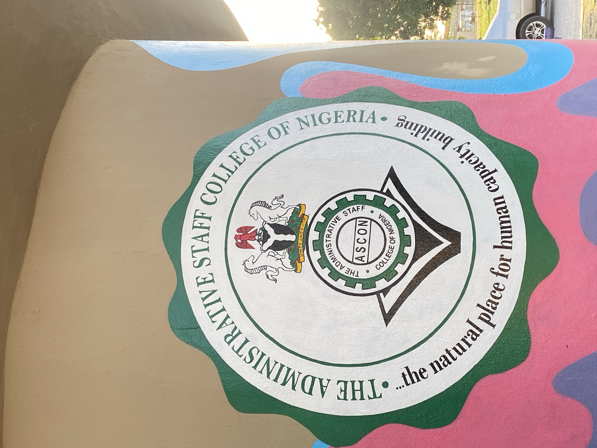 The image shows a close-up of a painted design featuring the logo of "The Administrative Staff College of Nigeria" (ASCON). The logo is circular, with a green scalloped border. Inside, there's a white circle containing the college's emblem, a black-and-white heraldic crest, and a gear-like design with the acronym "ASCON" in the center. The text "THE ADMINISTRATIVE STAFF COLLEGE OF NIGERIA" surrounds the upper half of the circle, and "...the natural place for human capacity building" is written around the lower half. The painted surface around the logo has a camouflage-like pattern with colors like brown, blue, pink, and purple. The location is Tawpo, Nigeria.