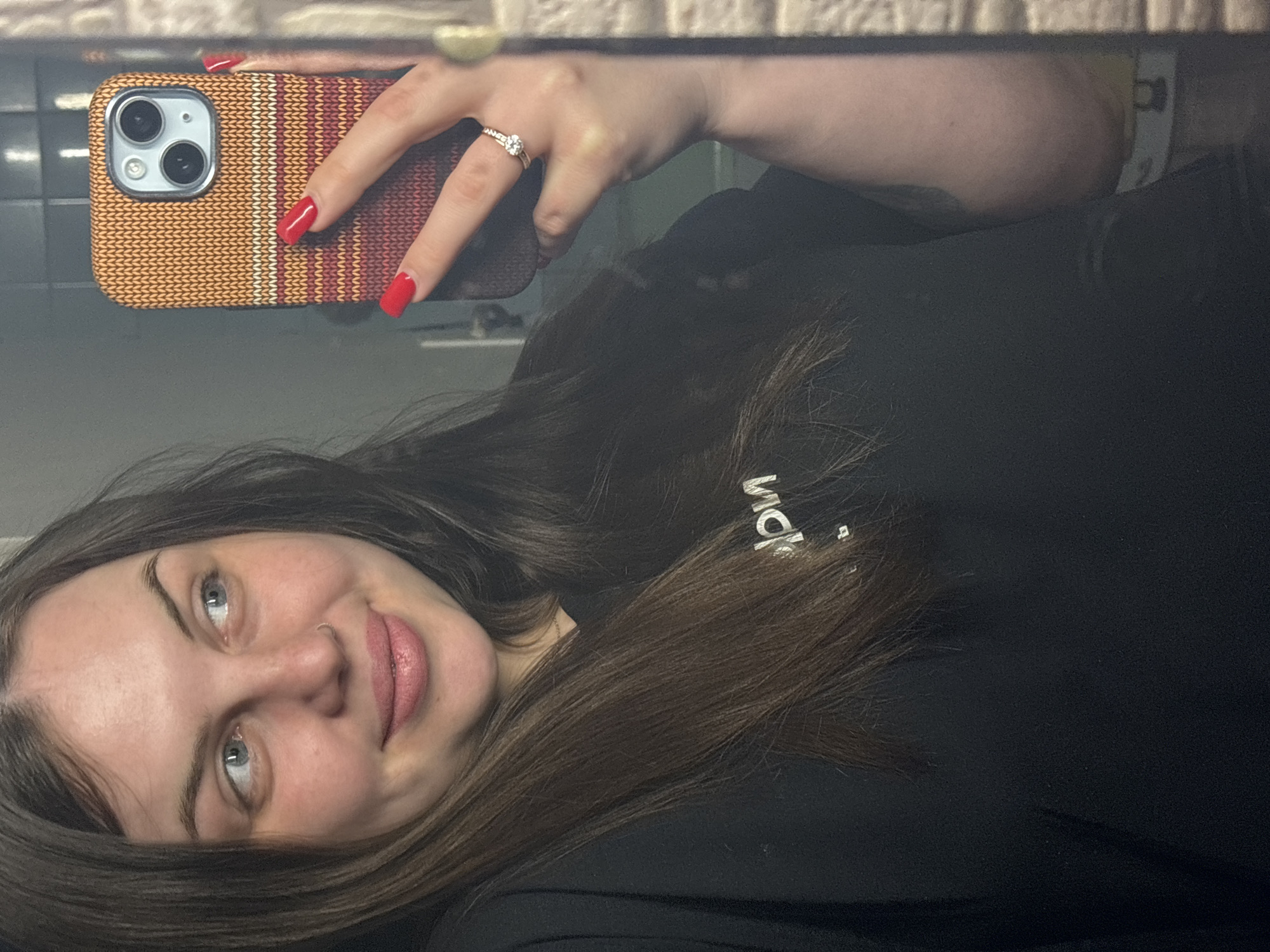 A woman is taking a selfie in a mirror. She has long brown hair, blue eyes, and is wearing a black shirt with white lettering. She is holding a smartphone with a patterned case, and her nails are painted red. She is wearing a ring on her left ring finger. The background includes a tiled wall. There is no information to deduce any location in the city of Chornomorskoe, Ukraine.