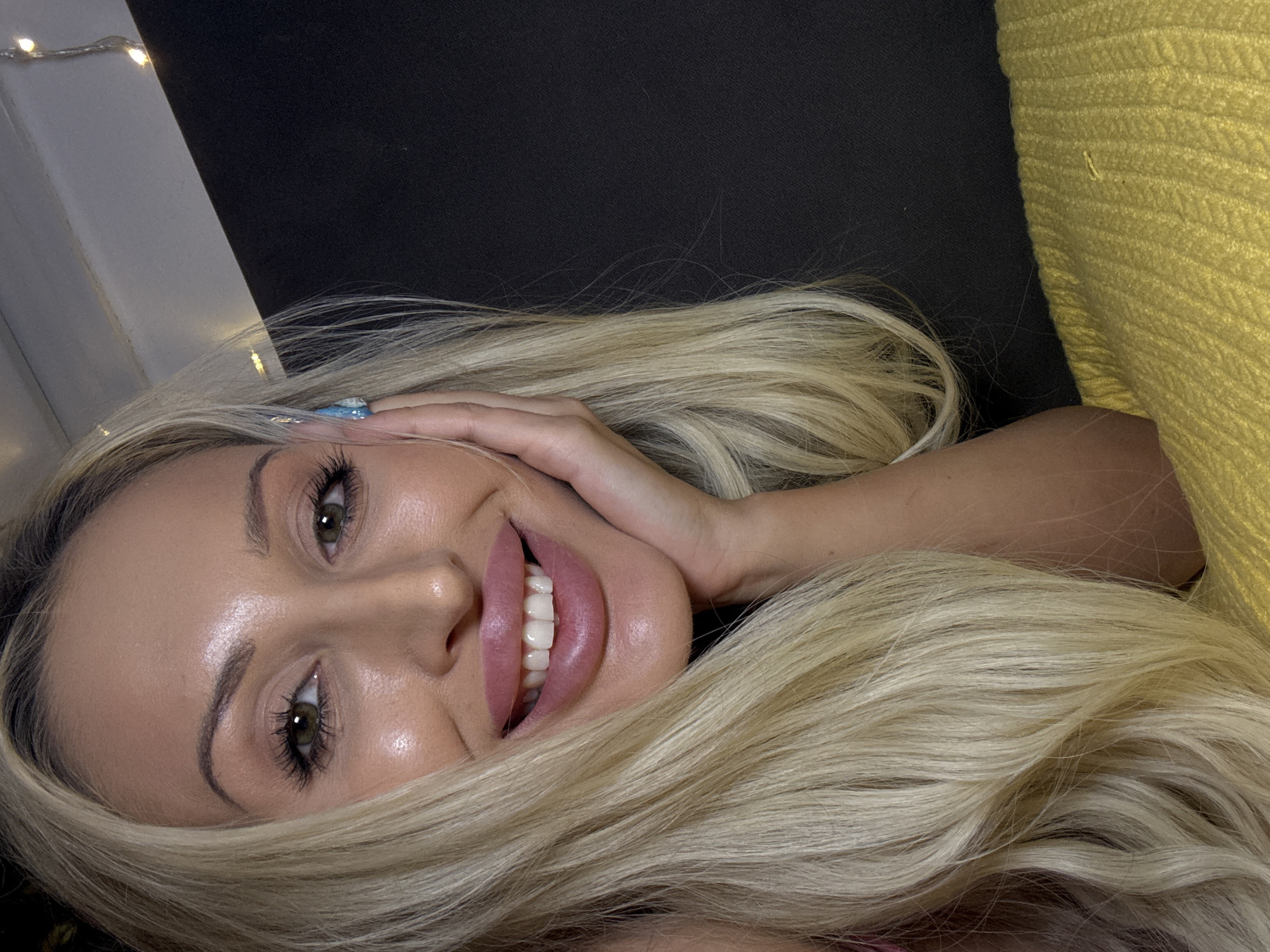 A young Caucasian woman with long blonde hair and green eyes smiles at the camera, resting her hand on her cheek. Her lips are painted pink, and her teeth are visible. She is wearing makeup, including eyeshadow and mascara, and has long, painted fingernails. The background is dark, with some decorative lights visible at the top. The woman is lying down on a textured yellow surface. The image appears to be a selfie.