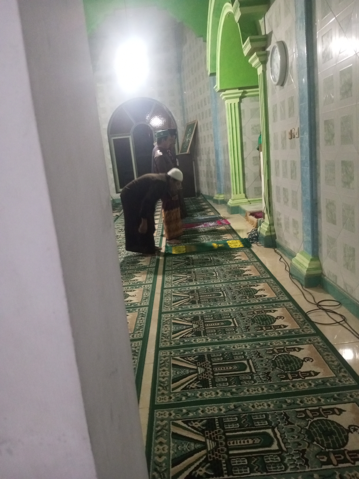 In Andongsili, Indonesia, a dimly lit interior space, possibly a mosque or prayer hall, is captured. The scene features a long, patterned green carpet laid out on the floor, with a repeating motif of mosque-like structures. In the foreground and midground, two individuals are partially visible, engaged in prayer or observing the prayer. One person, wearing dark clothing and a white cap, is bent over on the carpet. Another individual, wearing a patterned skirt and a darker top, is standing. The walls of the space are adorned with a grid pattern of tiles in light and blue colors. Green pillars with decorative molding accent the walls, and a large, arched entryway is visible in the background, illuminated by a bright light source. A circular clock is mounted on the tiled wall. Electrical cords are seen on the floor near the wall.